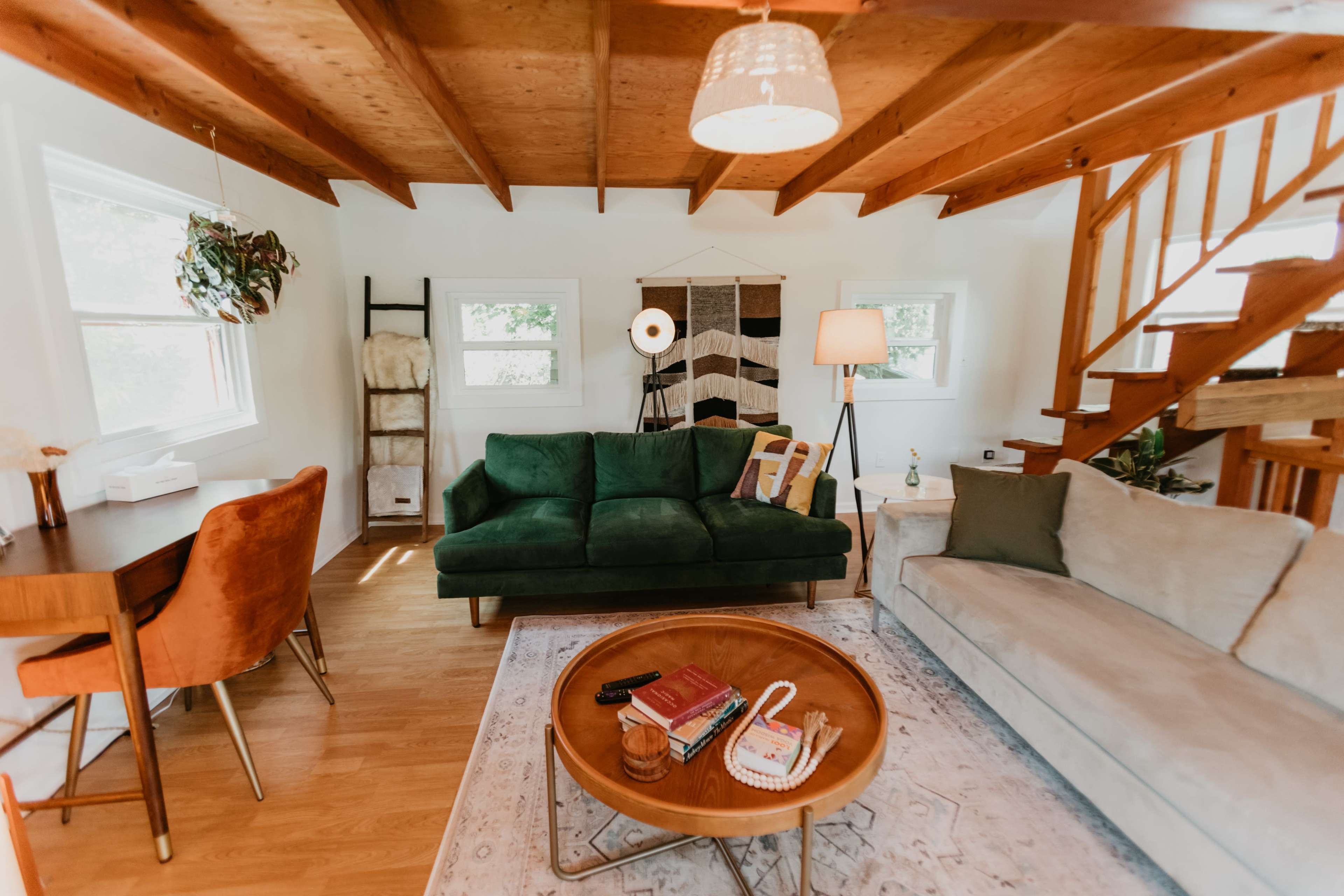 A cozy living room features a green sofa, a beige couch, a wooden coffee table, and a staircase, with large windows and decorative lighting.