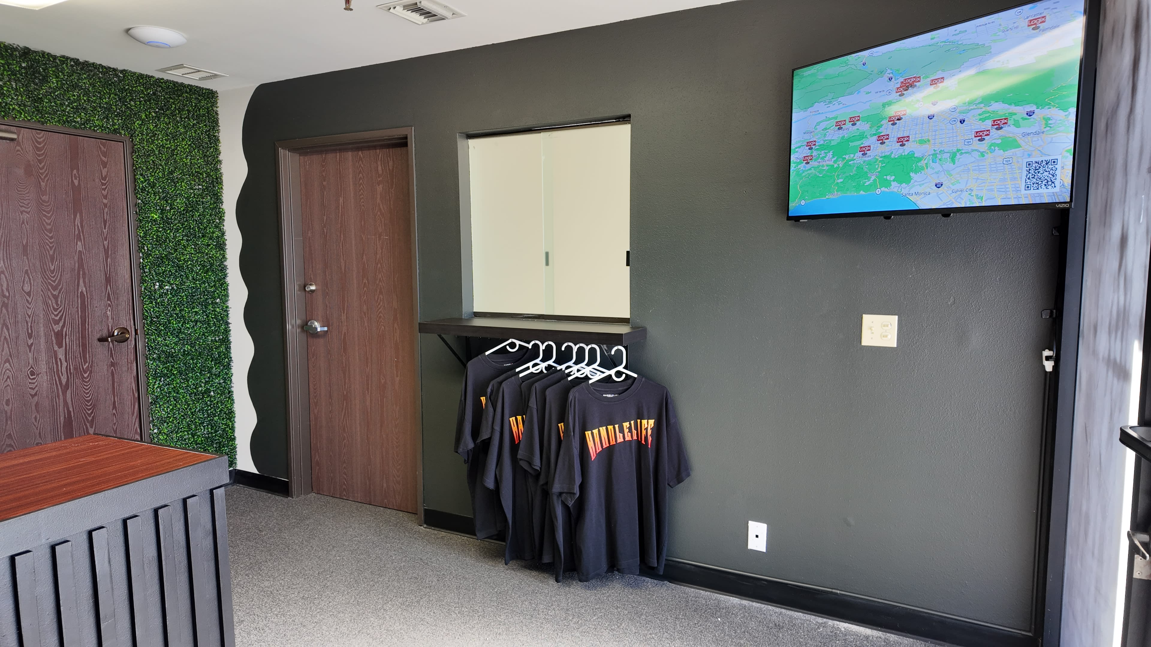The image shows a clean, modern interior space with a dark wall, a mirror, a television displaying a map, and hanging black t-shirts on a rack.