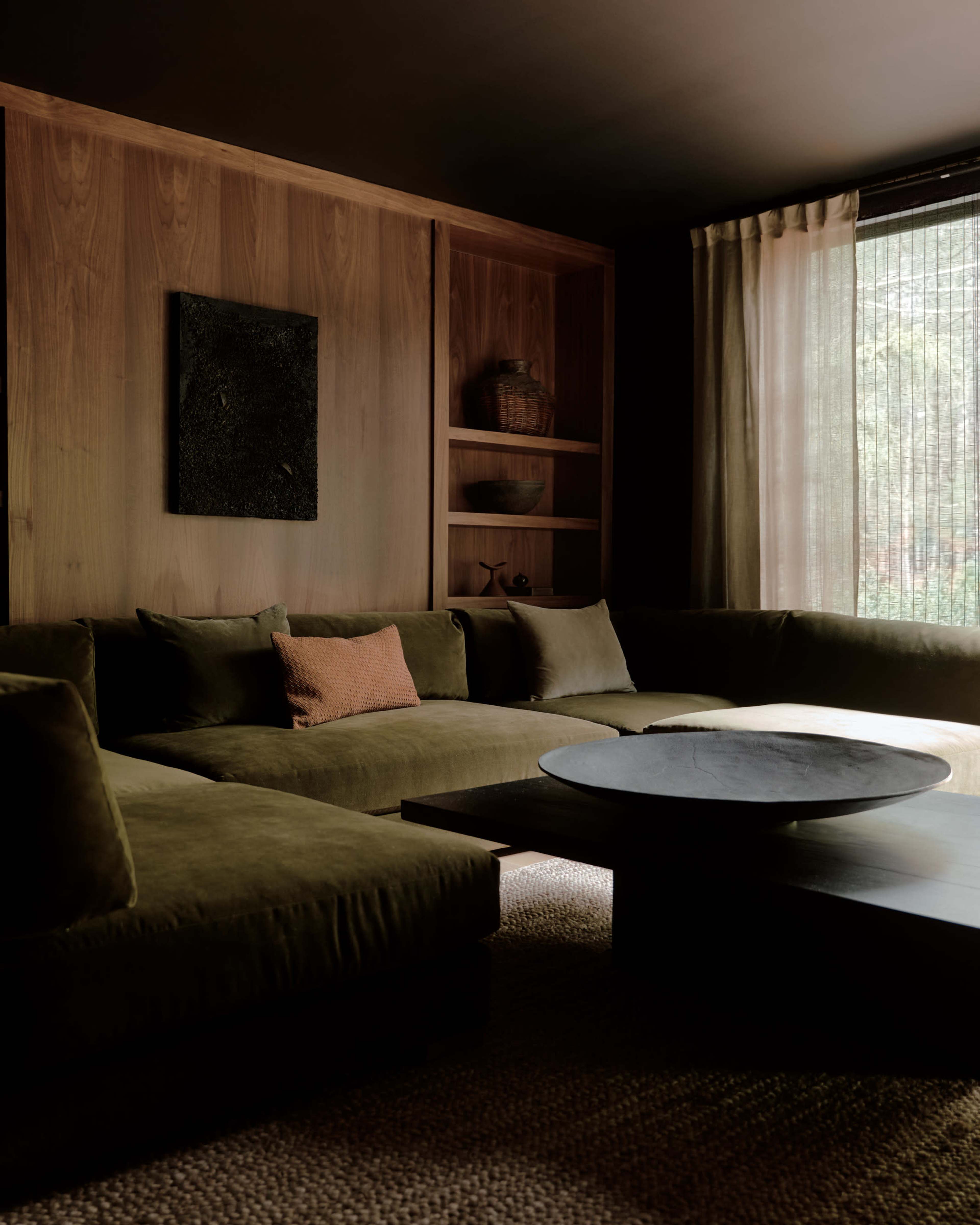 The image shows a cozy, dark living room featuring a large, L-shaped olive green sofa, a round black coffee table, and a wood-paneled wall with minimal decor.