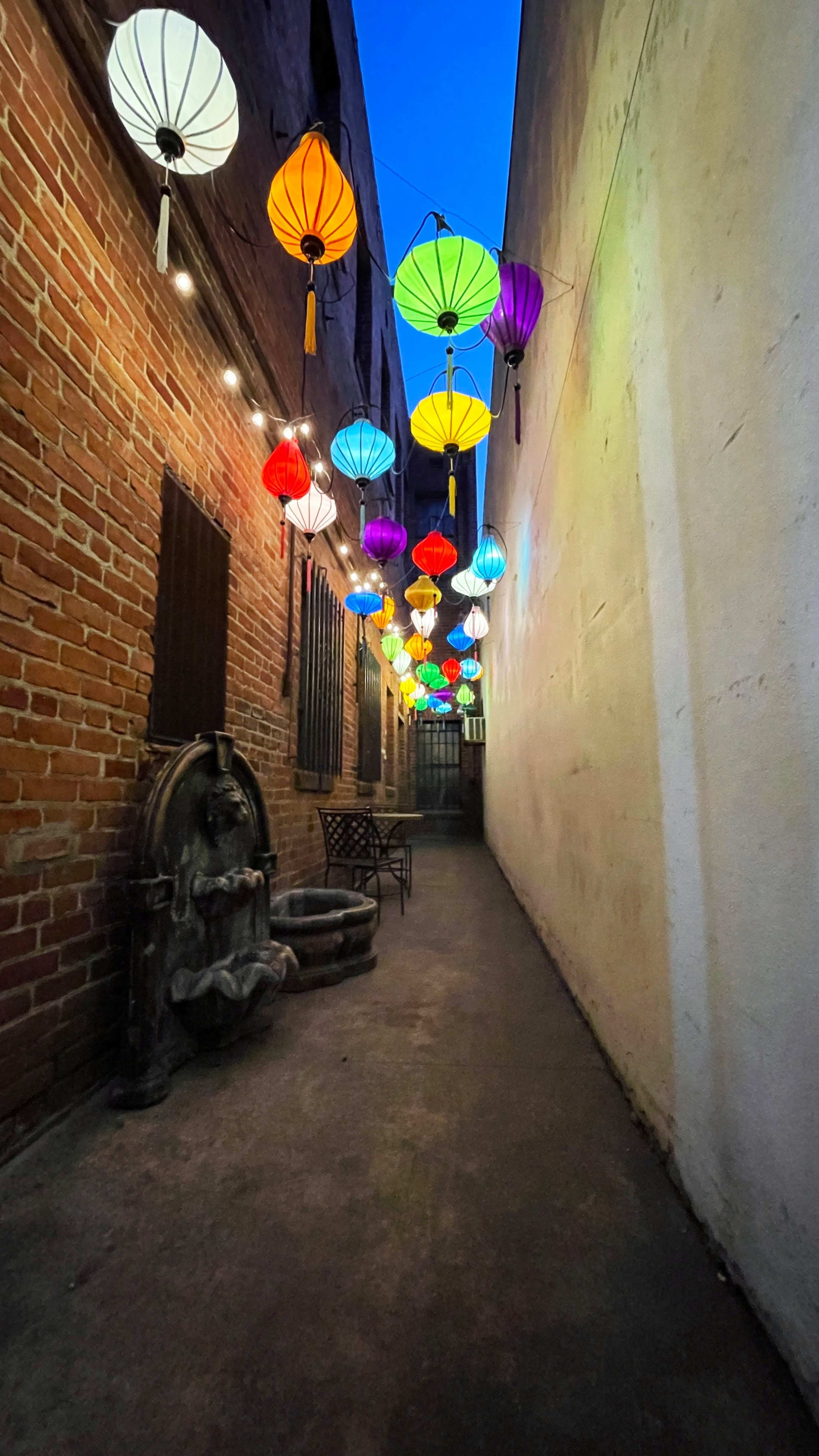 A narrow alleyway illuminated by colorful lanterns hanging above, with brick walls on one side and a stone fountain on the other.