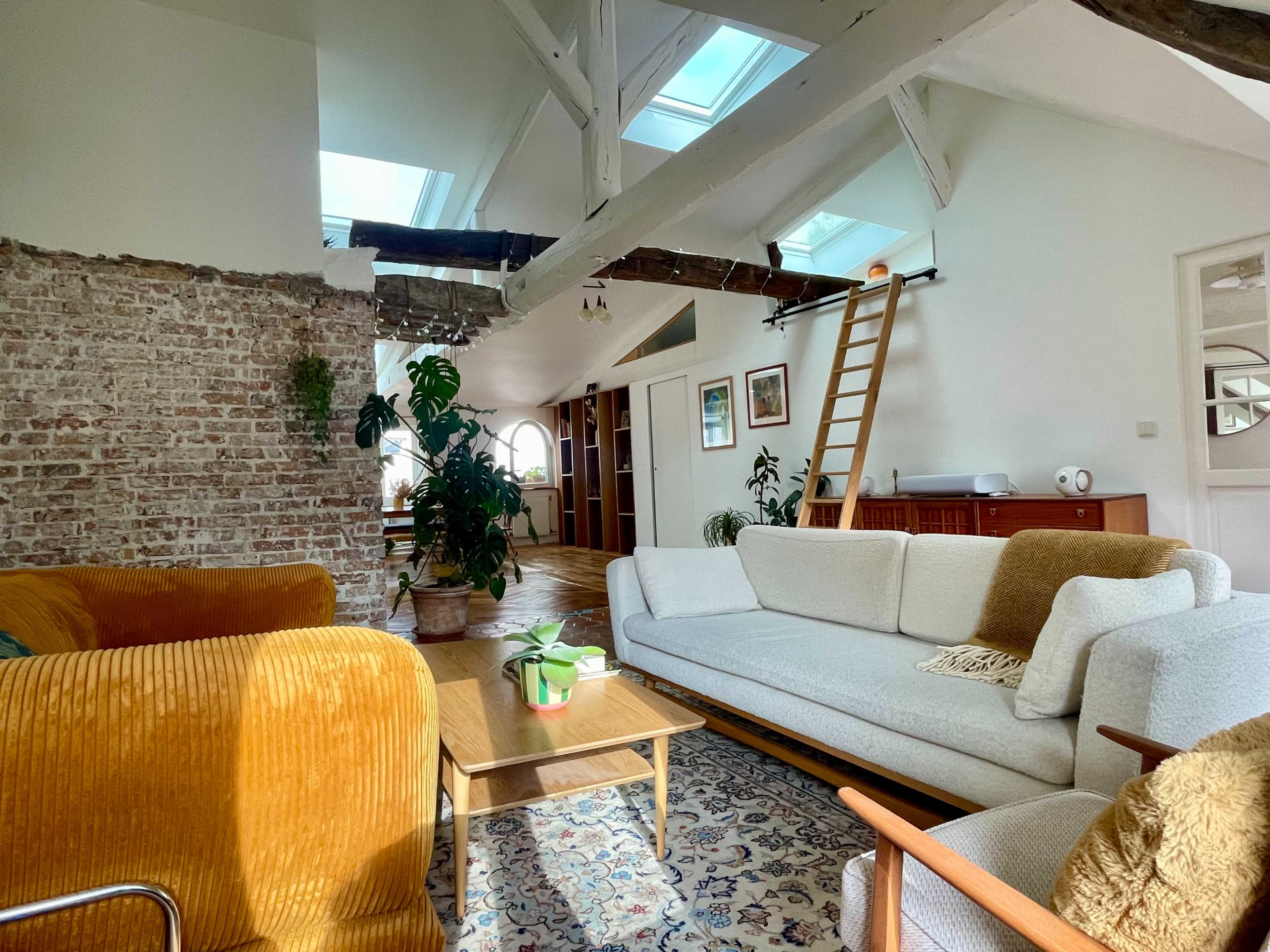 The image shows a spacious, well-lit living area featuring a mix of modern and rustic design elements, including a white couch, wooden furniture, and a climbing ladder leading to an upper level.
