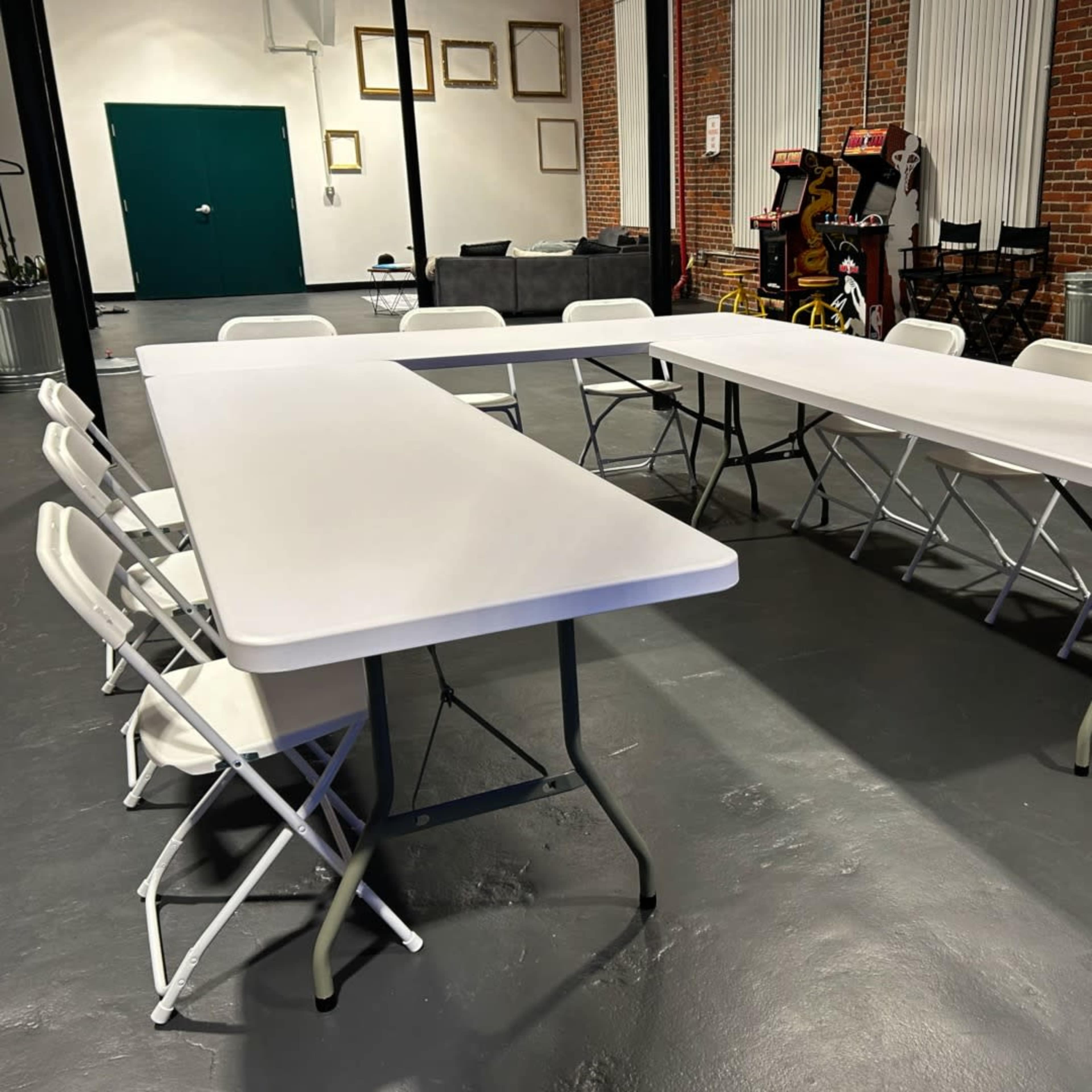 A set of white folding tables arranged in a rectangular shape with several folding chairs around them in a brick-walled room.