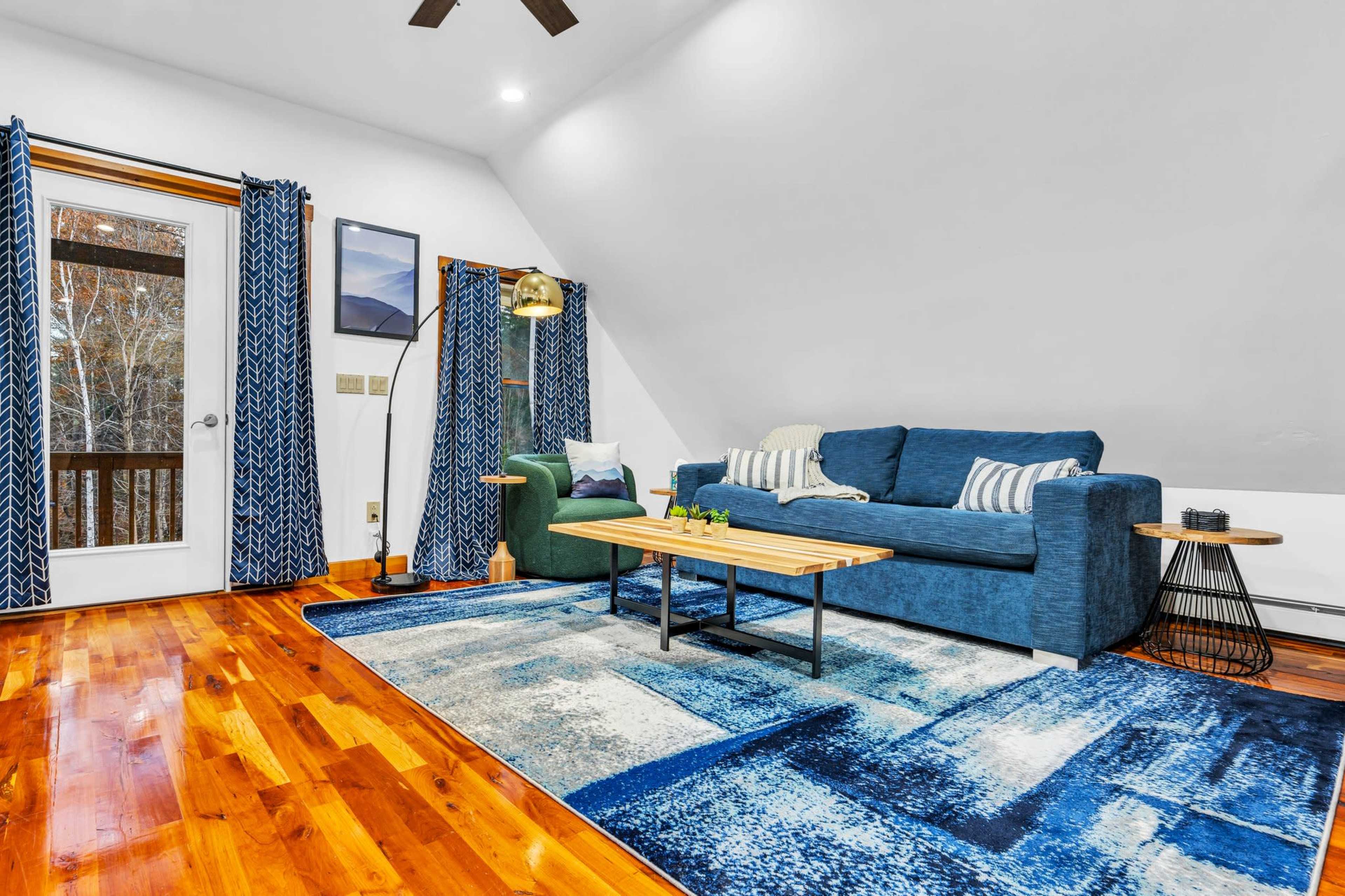 A cozy living room with a blue sofa, green chair, wooden coffee table, and large area rug, featuring a door leading to a balcony and large windows with patterned curtains.