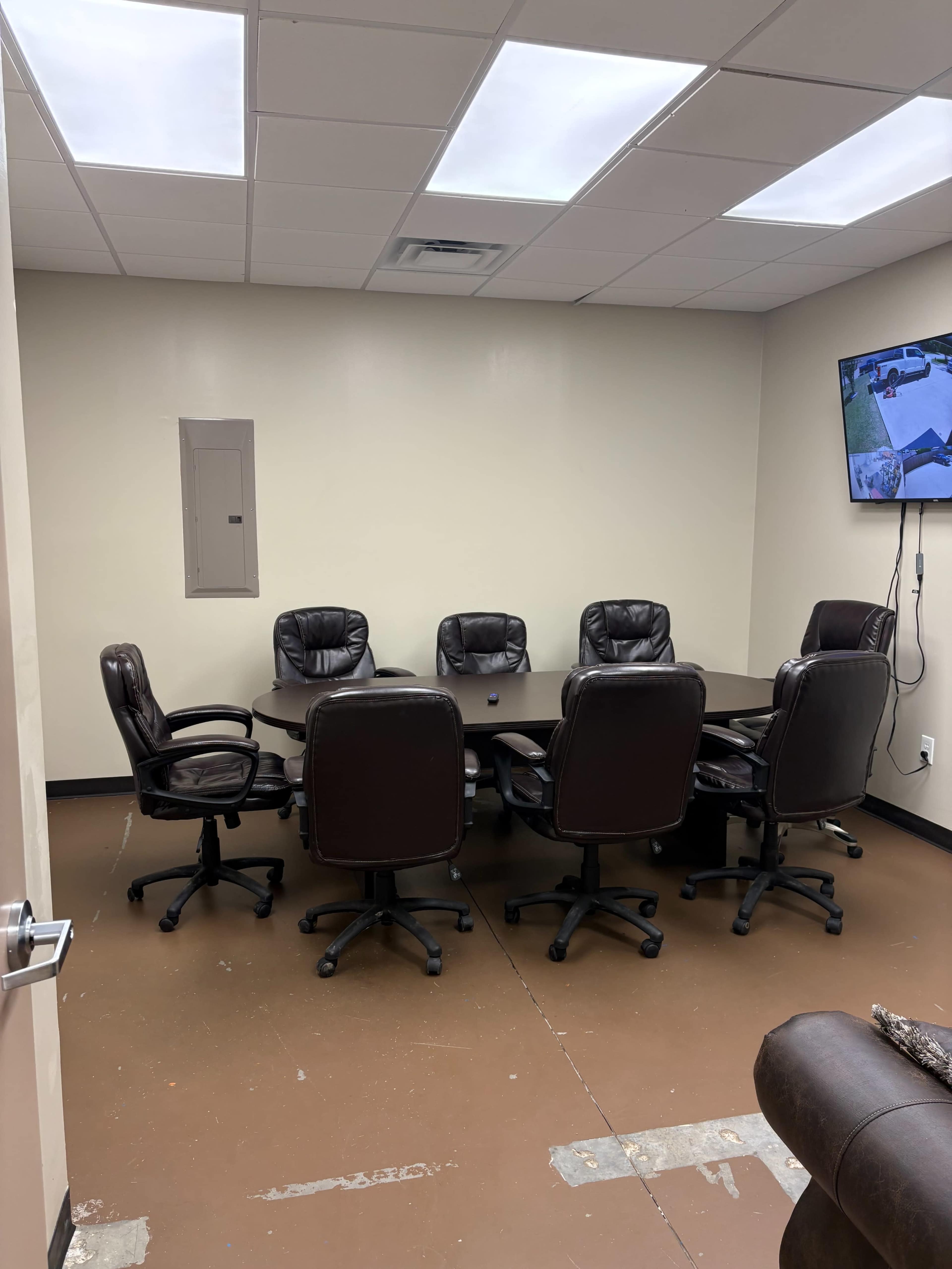 A conference room is equipped with a large table surrounded by eight leather office chairs and features a wall-mounted TV.