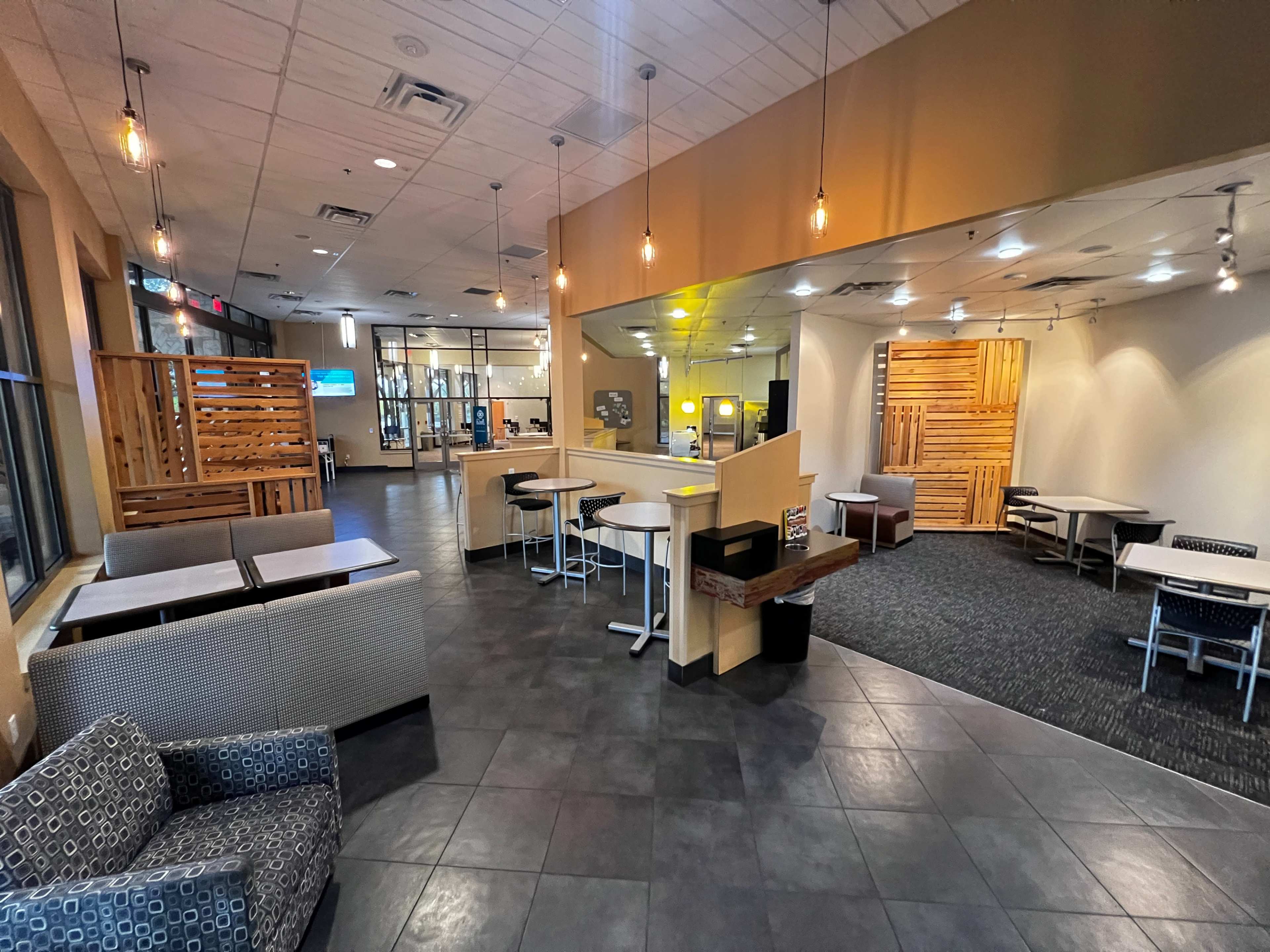 The image shows a modern, open interior of a public space with tables and seating arranged in a contemporary style, featuring wooden accents and a mixture of lighting.