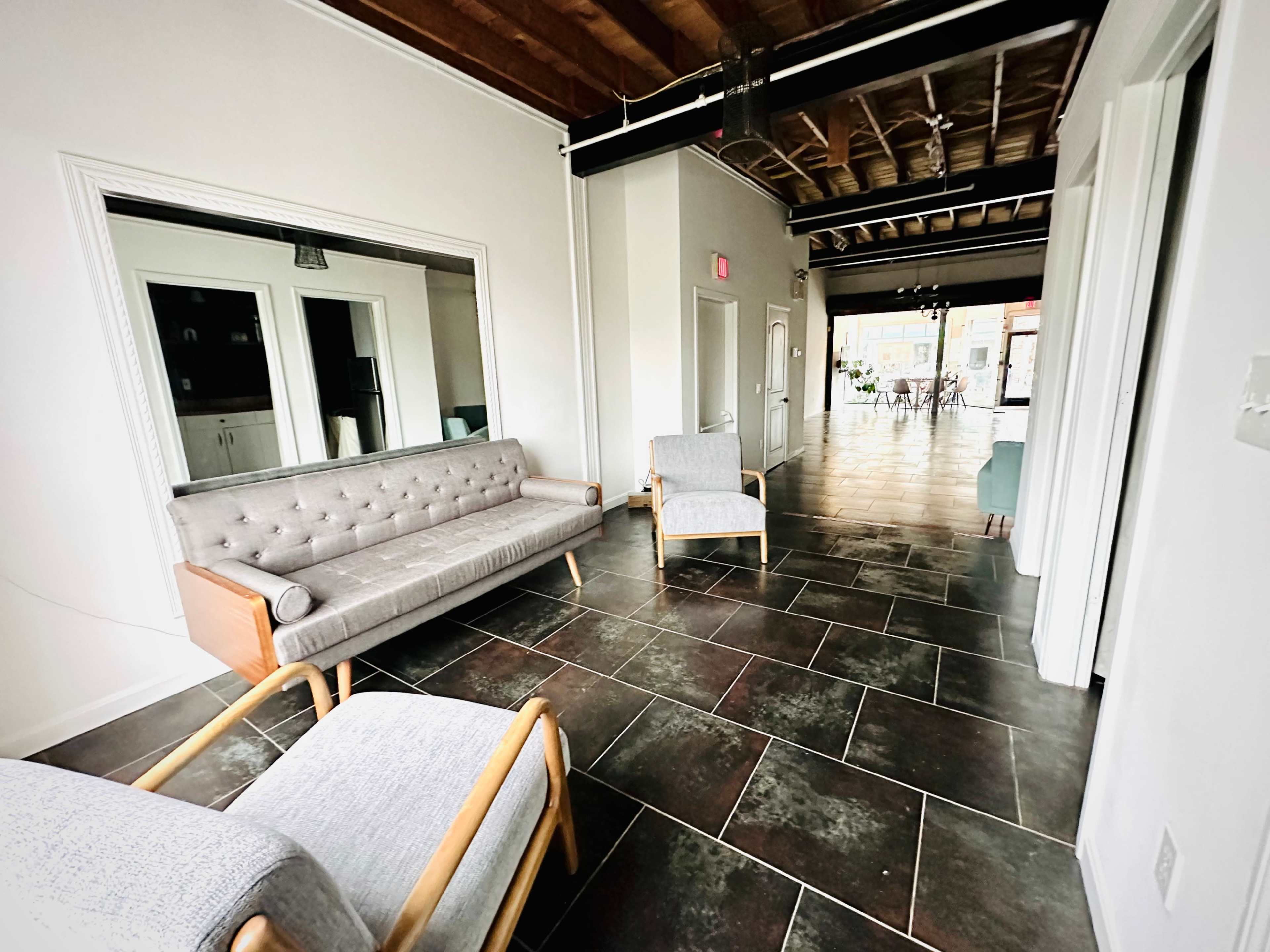 A spacious interior with a gray tufted sofa and two armchairs positioned on dark tiled flooring, leading towards an open area with large glass doors.