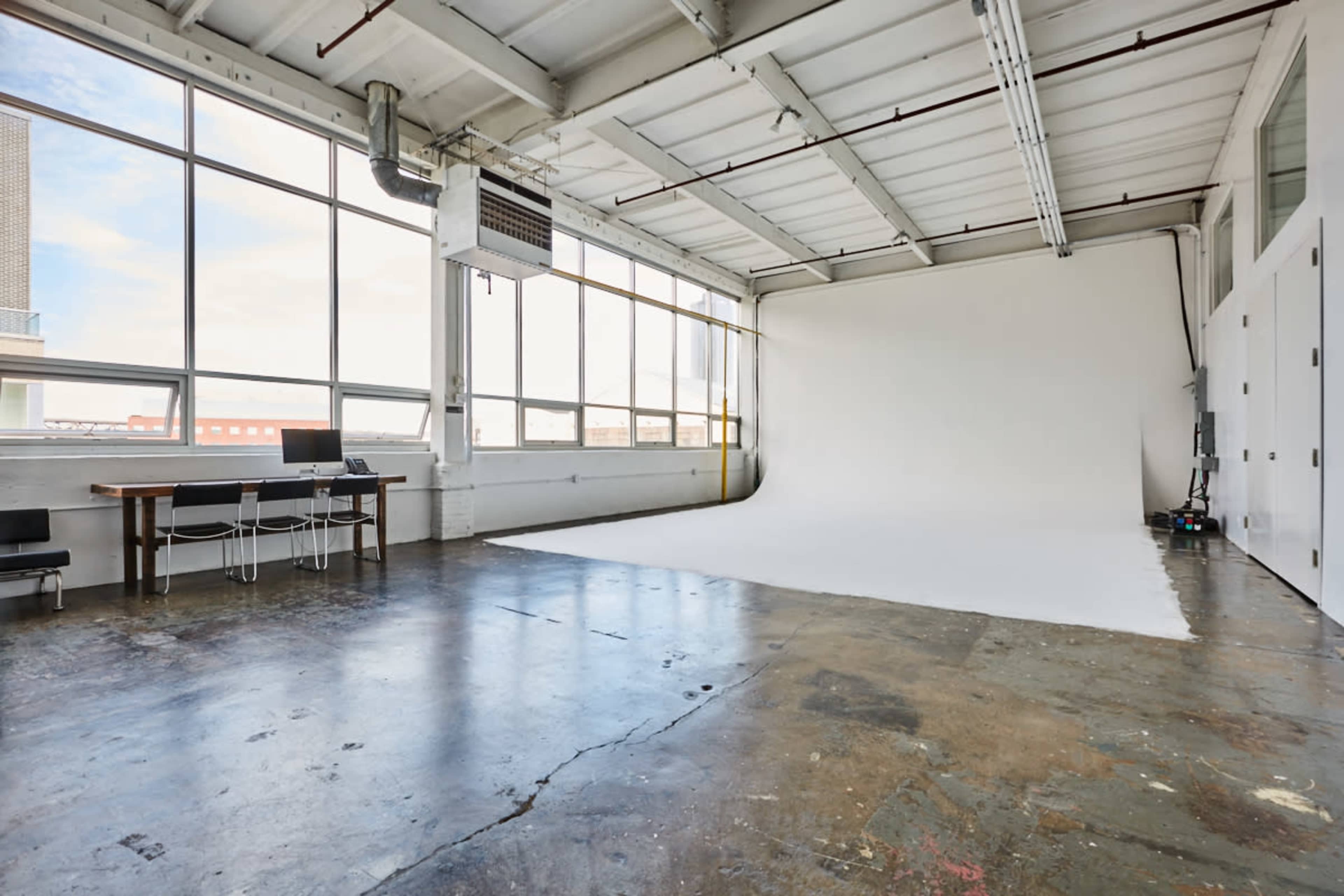 The image shows a spacious, empty photography studio with large windows, a white backdrop, and a wooden table with chairs.