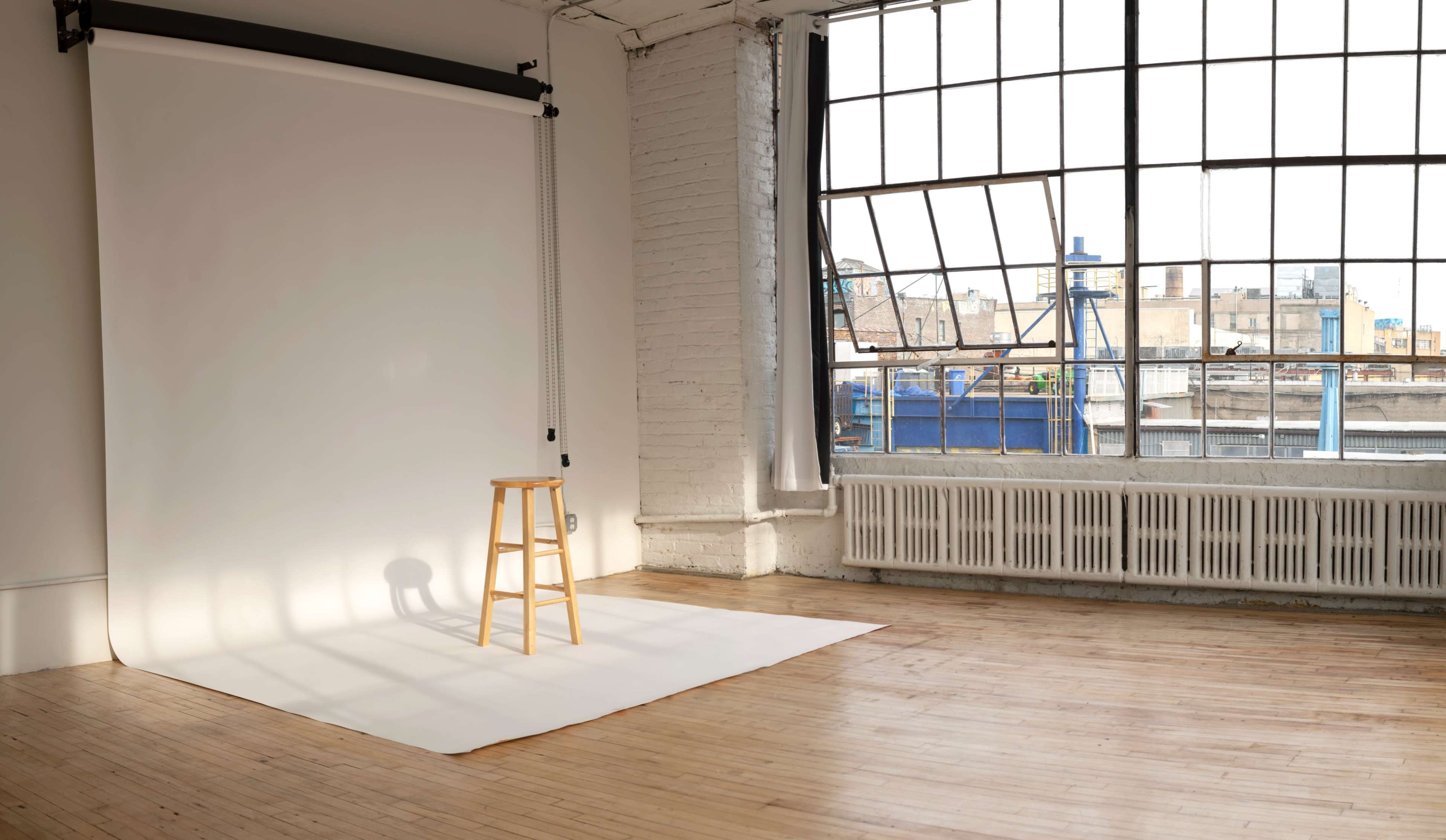 A wooden stool sits on a white backdrop beneath a large window in a well-lit studio space.