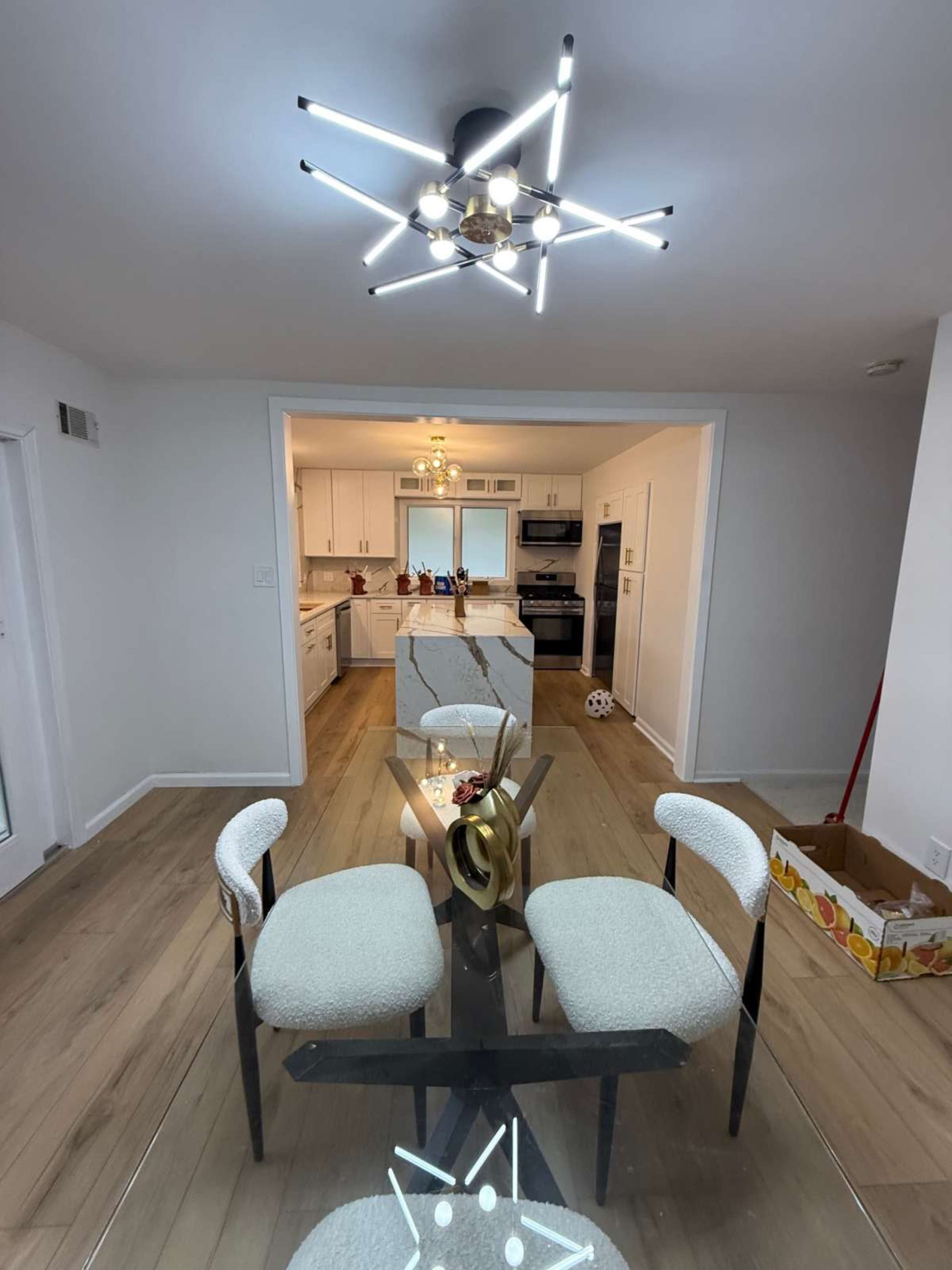 A modern dining area features a geometric ceiling light above a glass table surrounded by four upholstered chairs, with an adjoining kitchen visible in the background.