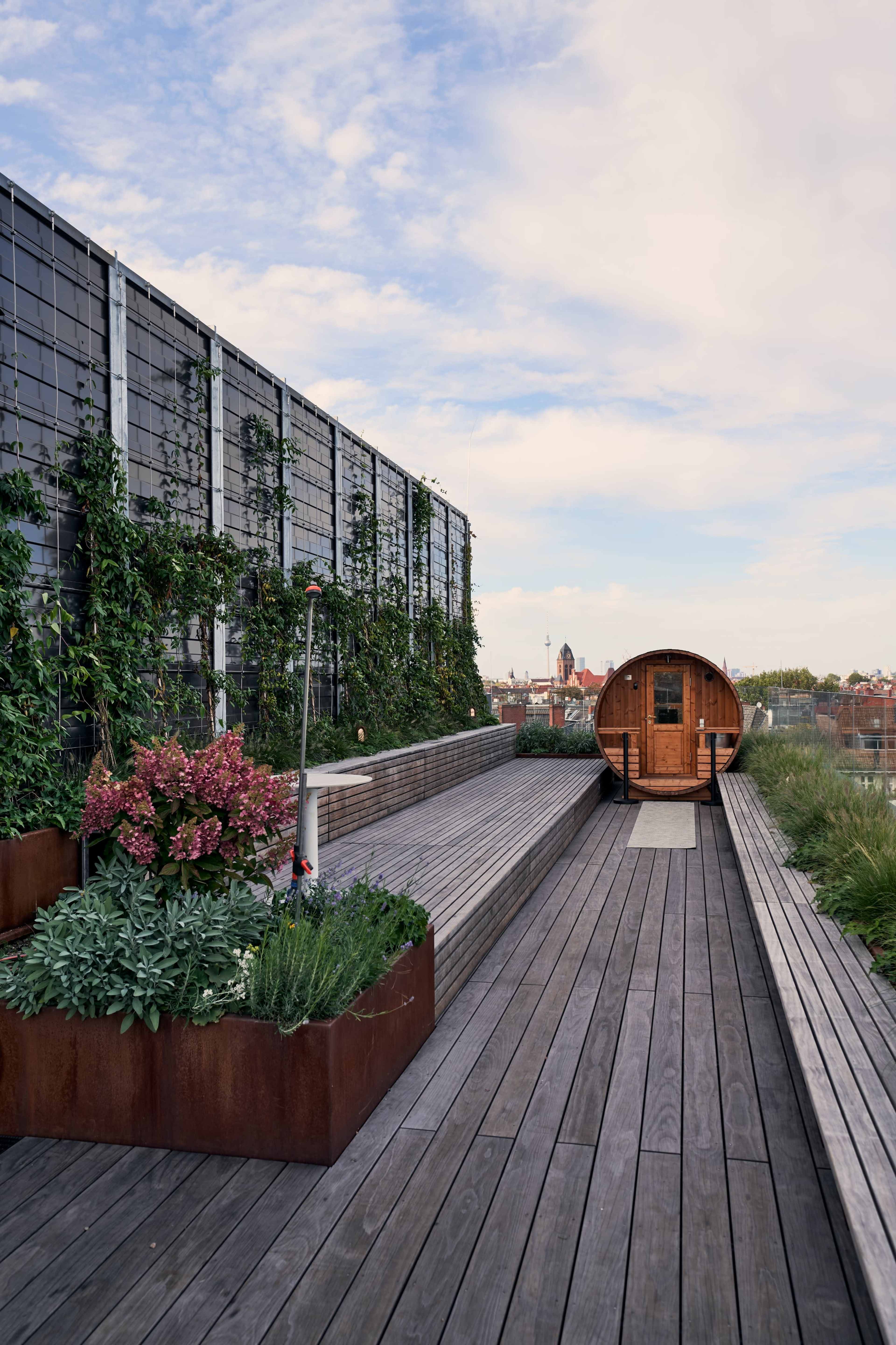 The image shows a modern rooftop space featuring wooden decking, raised planters with greenery, and a round wooden structure at one end against a backdrop of a blue sky.