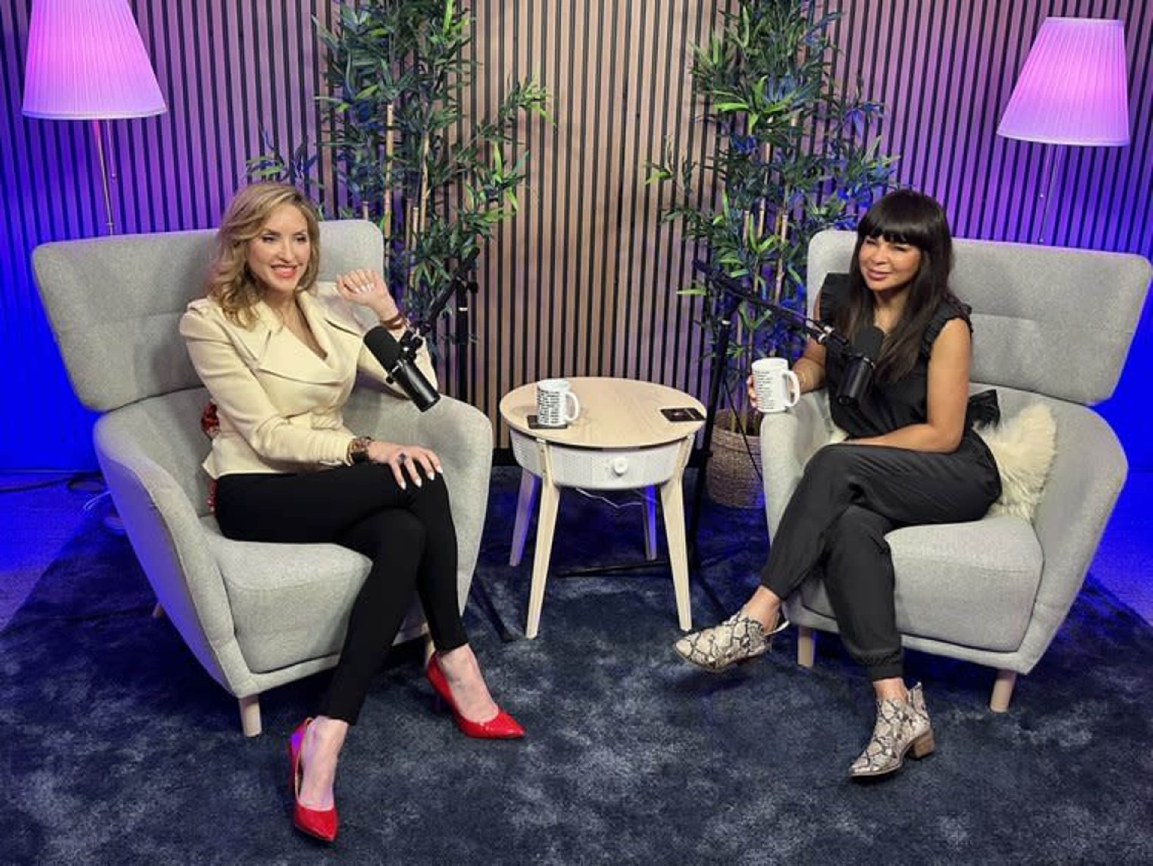 Two women sit in stylish armchairs in a modern studio, each holding a coffee mug and microphones are positioned near them.