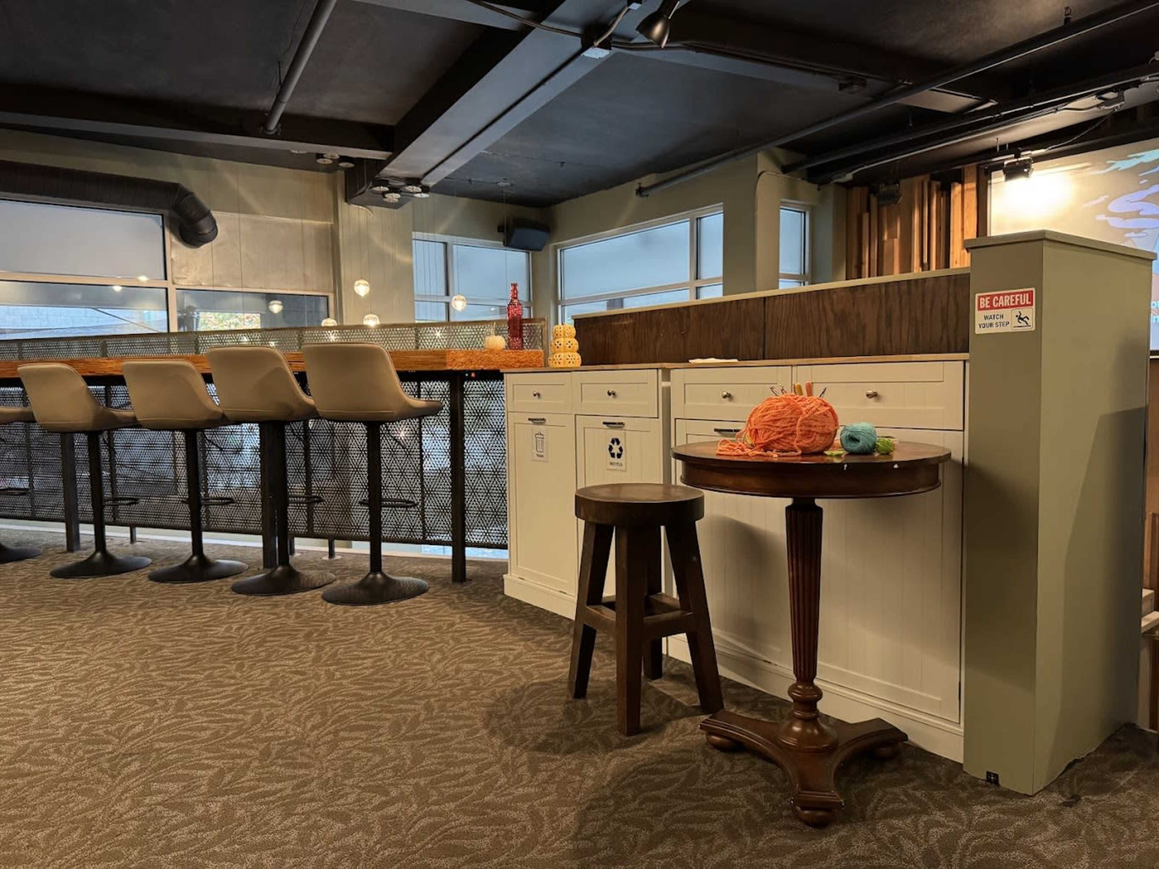 The image shows a modern bar area with a wooden counter, high stools, and a small round table featuring a spool of orange yarn and a toy fish.