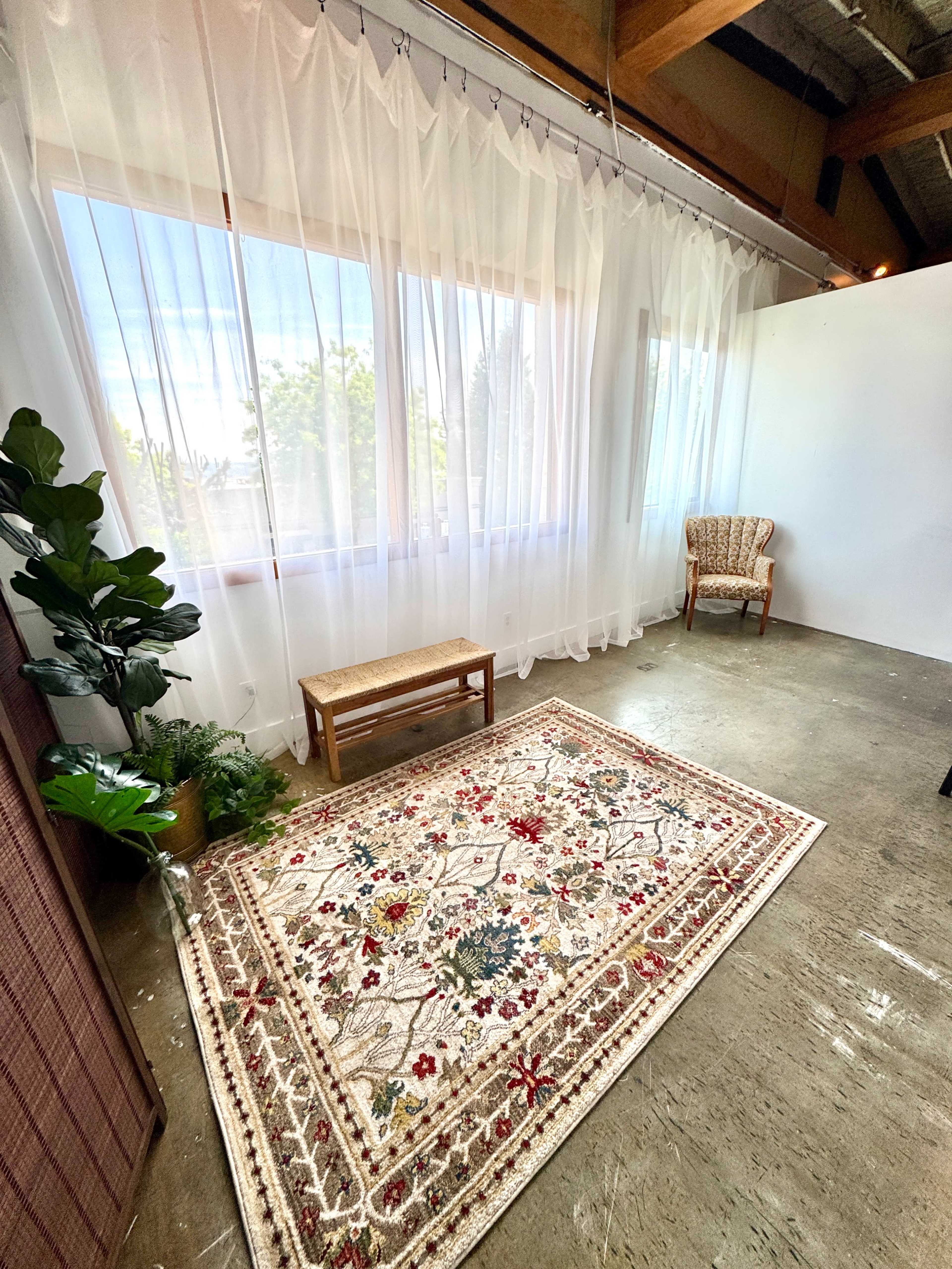 The image shows a well-lit room featuring large windows covered with sheer curtains, a decorative rug on the floor, a bench, a patterned chair, and a potted plant in the corner.