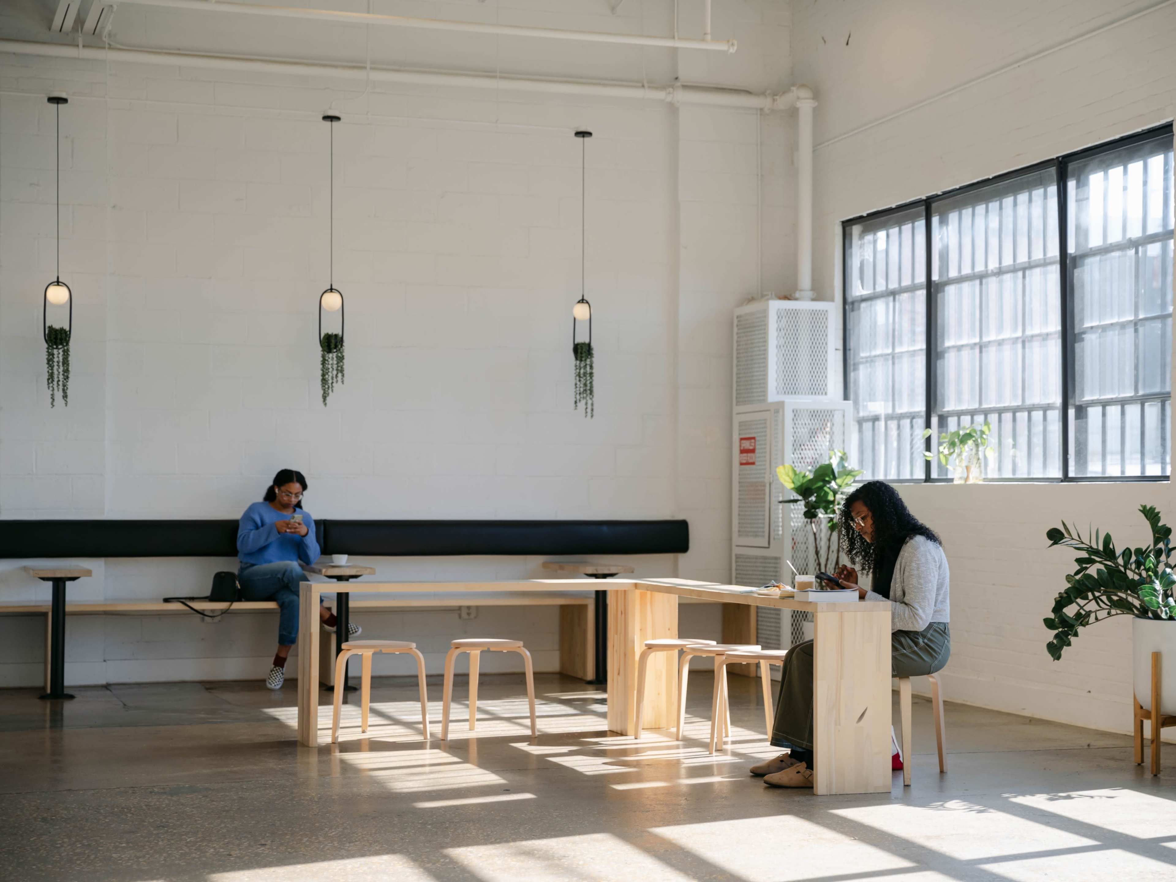 Two individuals are seated in a bright, minimalist café setting, each absorbed in their devices at a wooden table surrounded by plants.
