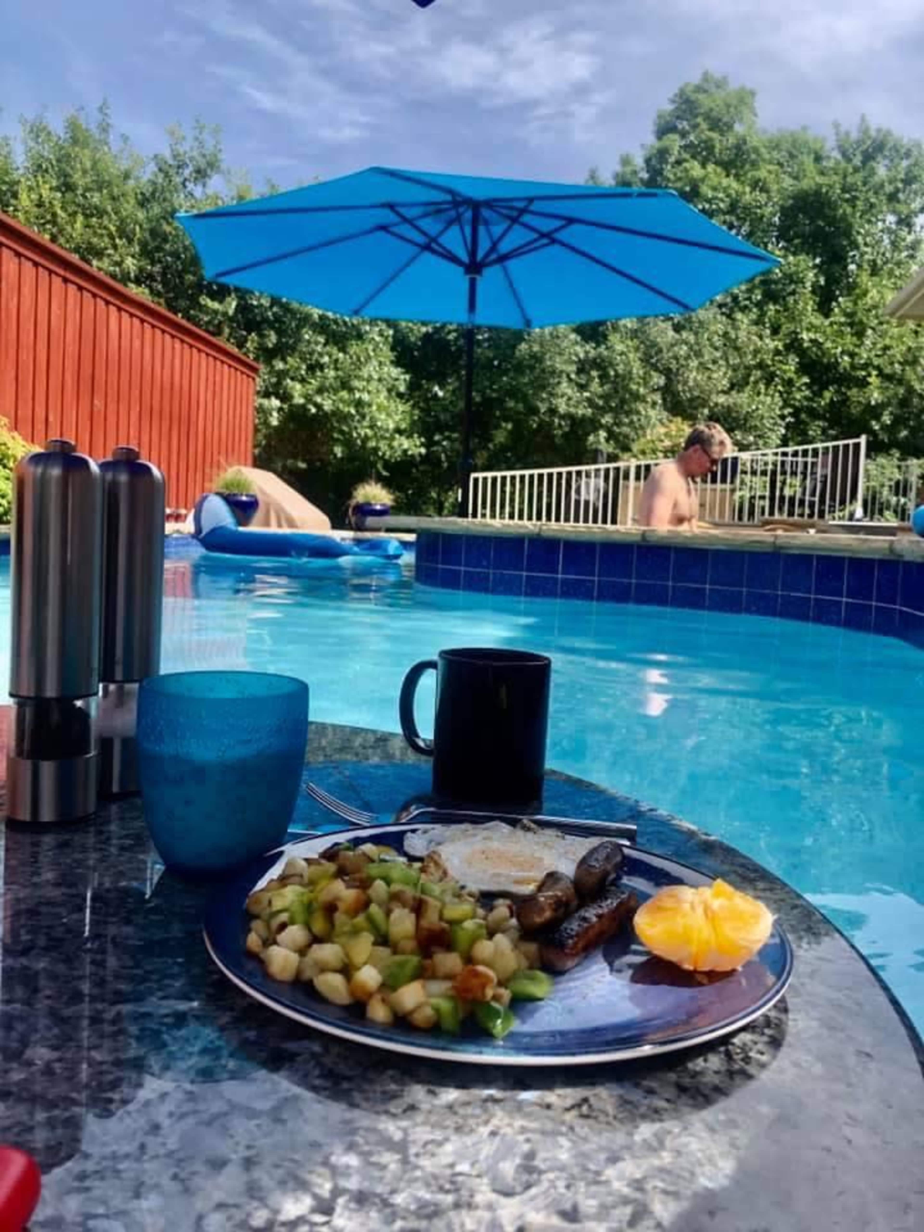 A plate of breakfast food sits on a table by a swimming pool, with an umbrella overhead and people relaxing in the water.