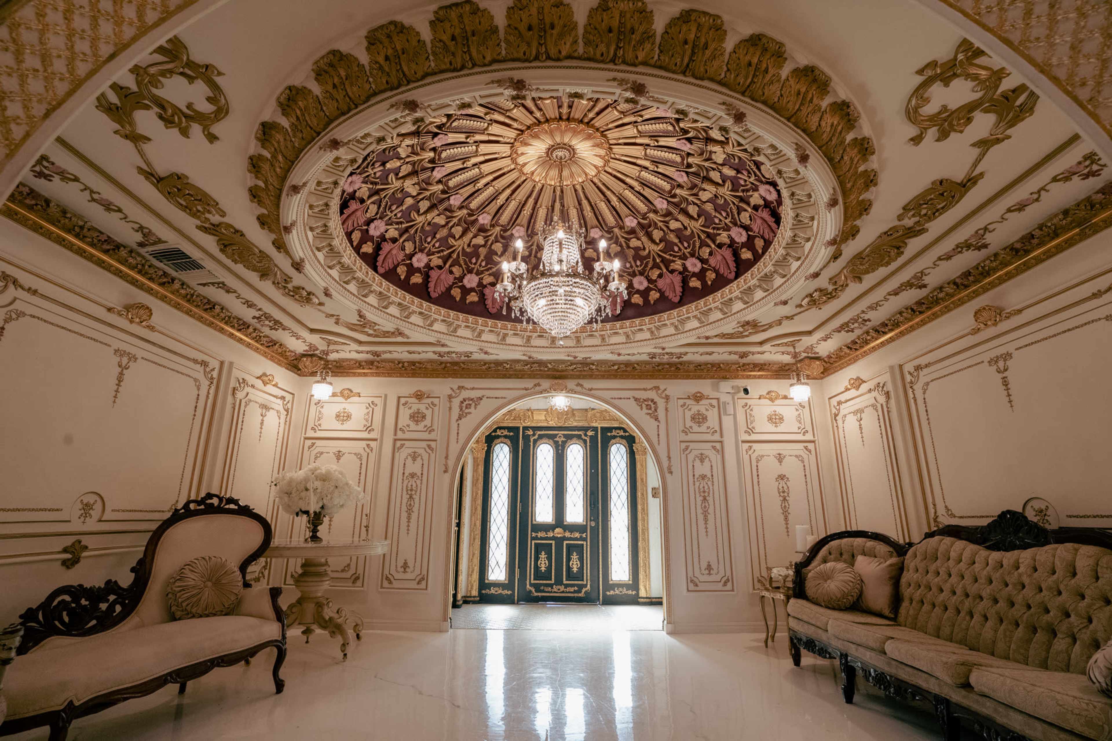 The image shows an ornate foyer with a chandelier, detailed ceiling, and ornate furniture, leading to a set of double doors.