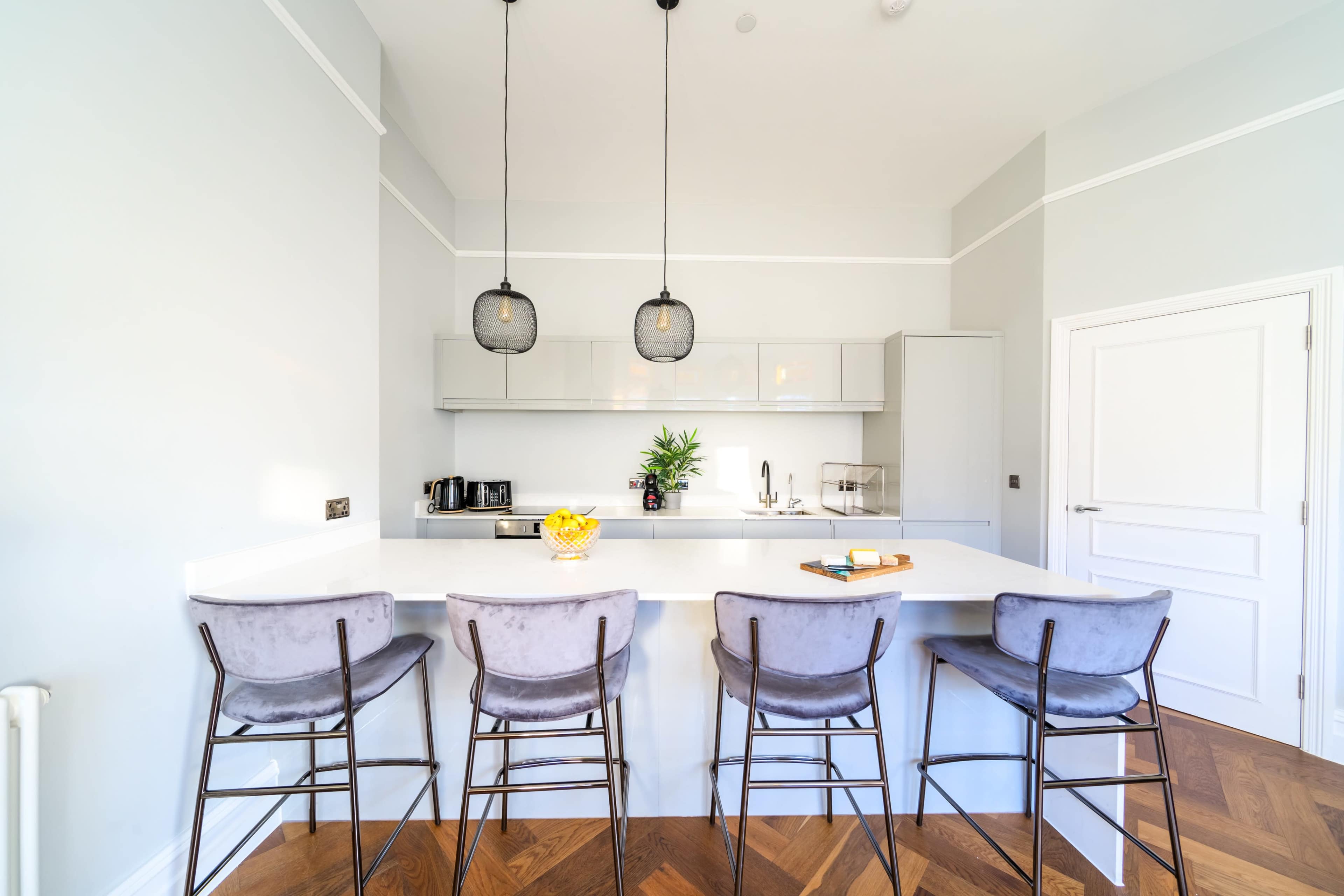 A modern kitchen features a white island countertop with four barstools, pendant lights hanging above, and minimalist cabinetry against a light gray wall.