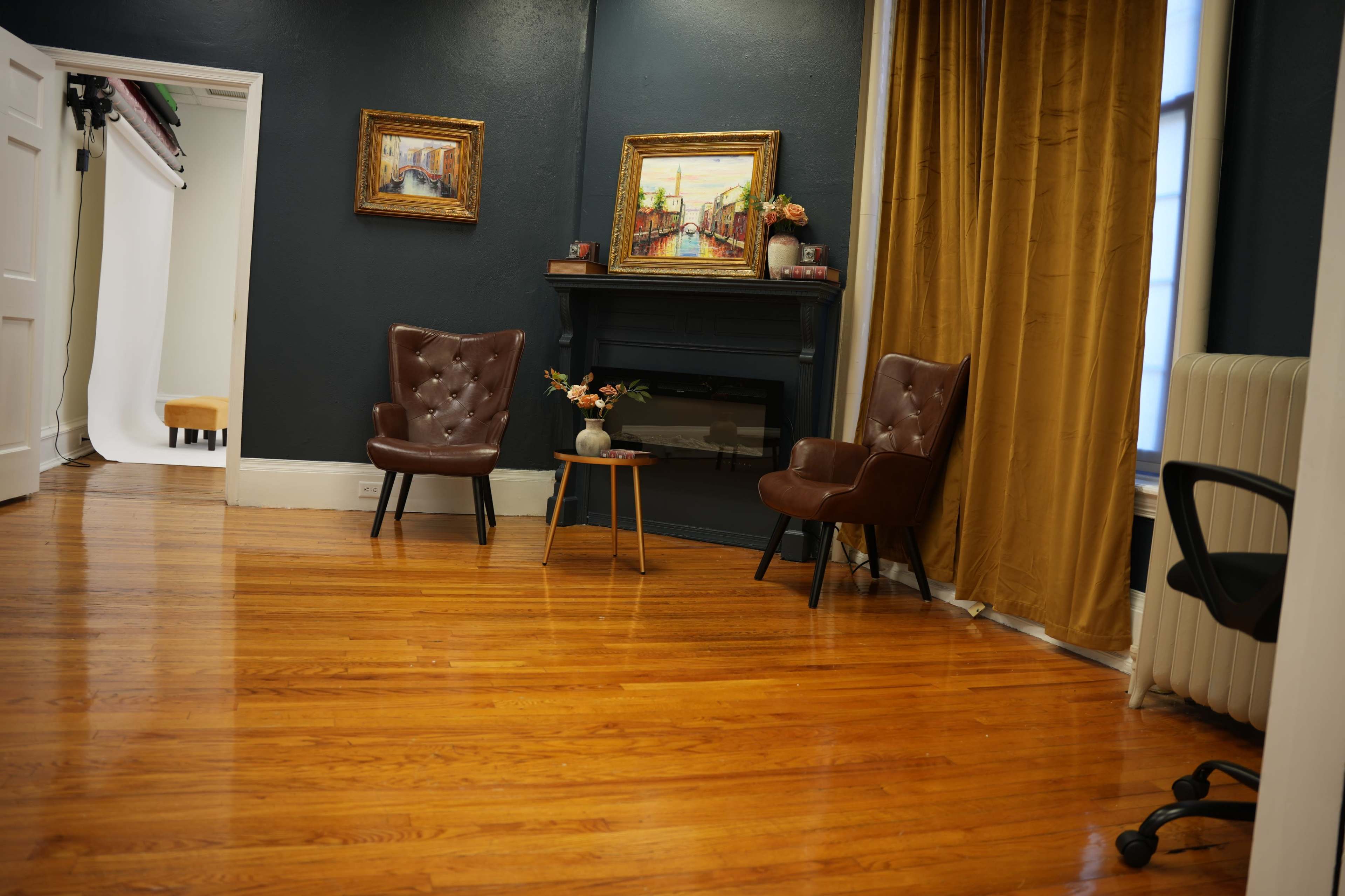 A room with dark walls, featuring two brown armchairs, a small table, and a decorative fireplace beside a window with yellow curtains.