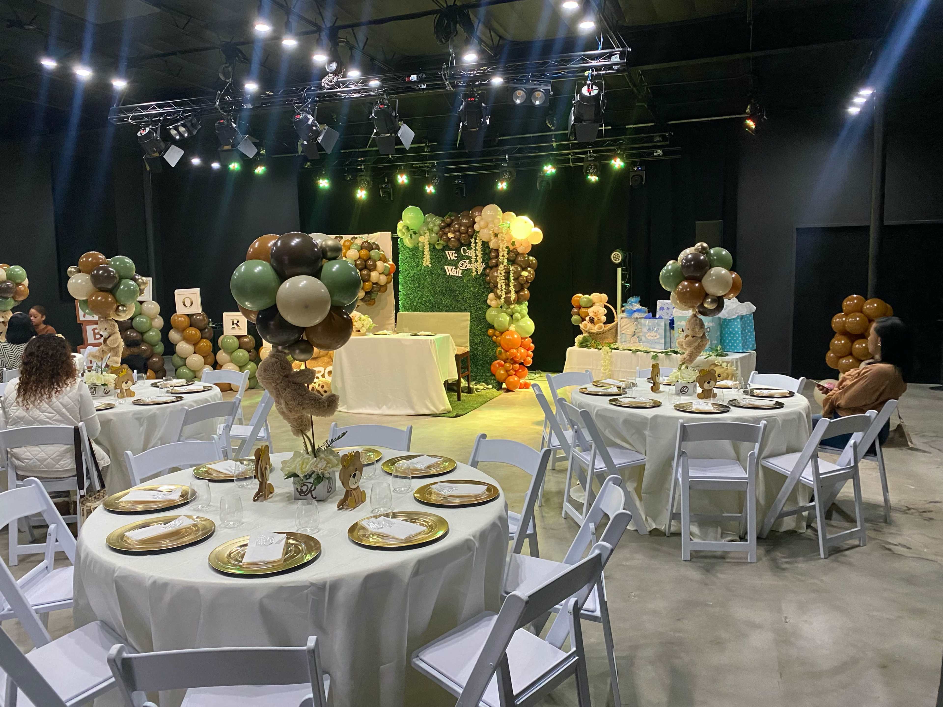 The image shows a decorated event space with round tables set for a gathering, featuring balloon displays and a backdrop adorned with greenery and decorative elements.