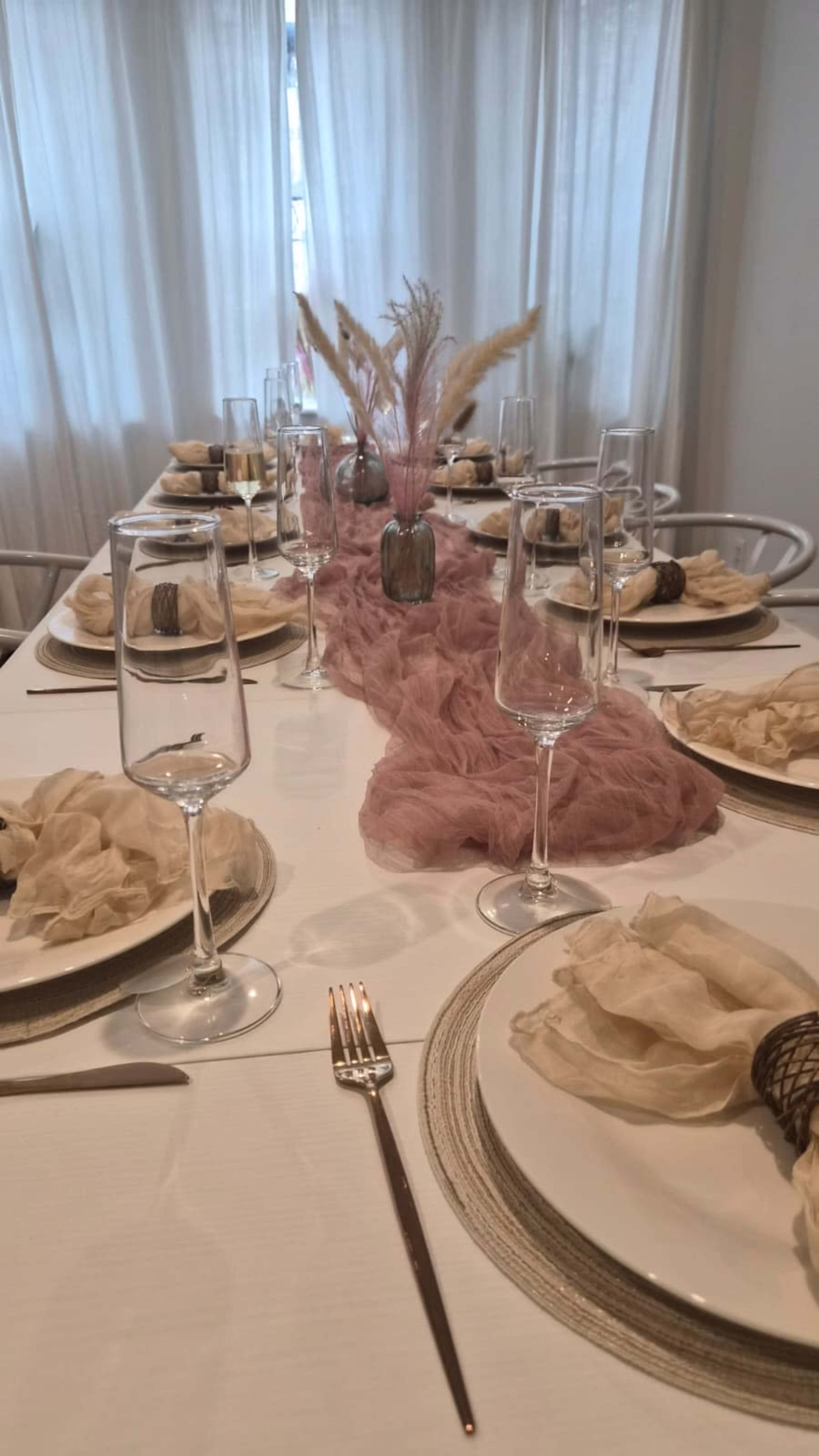 A long dining table is elegantly set with plates, glasses, and decorative elements, featuring a soft pink runner and floral arrangements in the center.