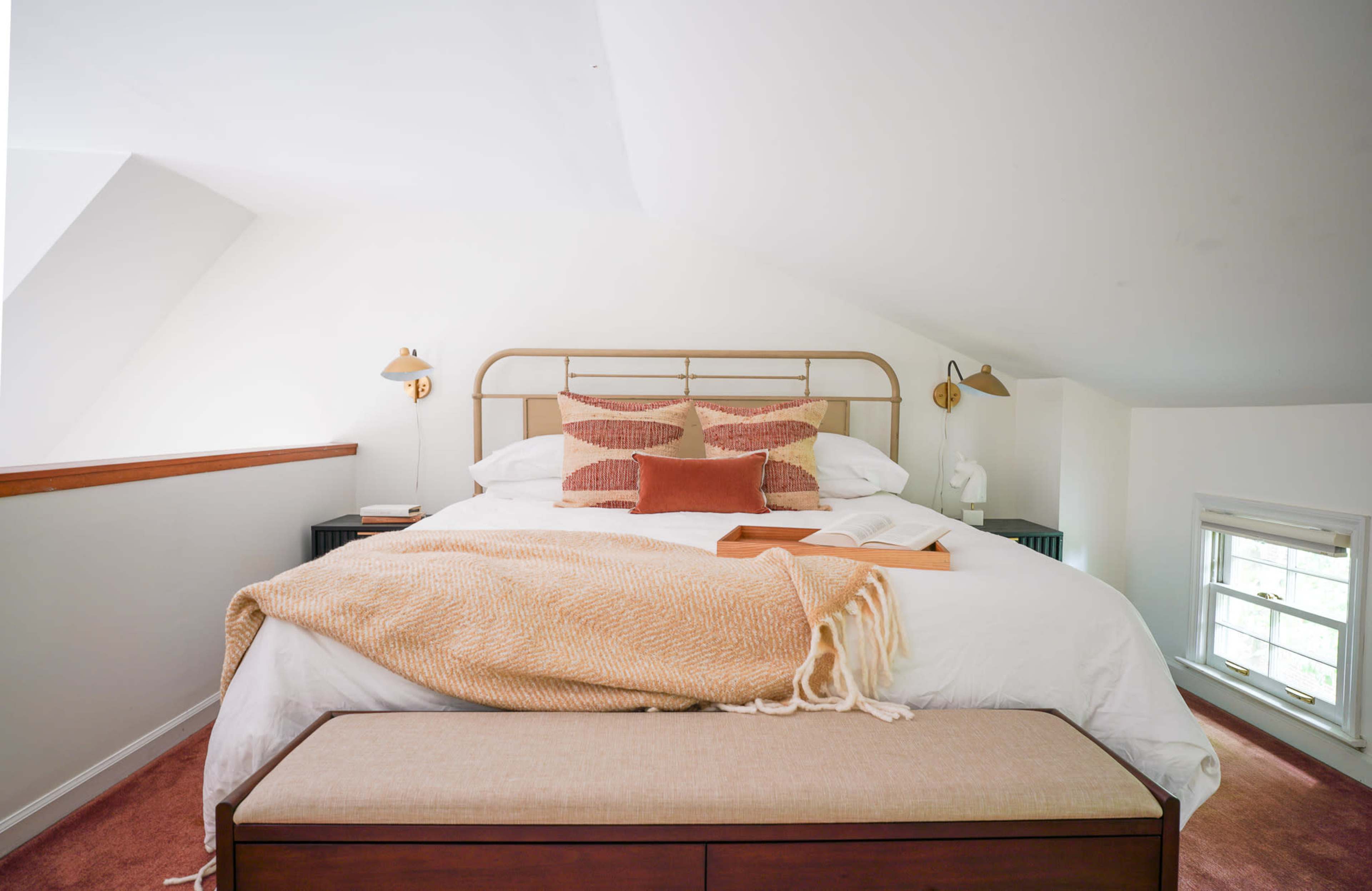 A neatly made bed with a patterned throw blanket and decorative pillows is positioned in a bright, sloped-ceiling bedroom.