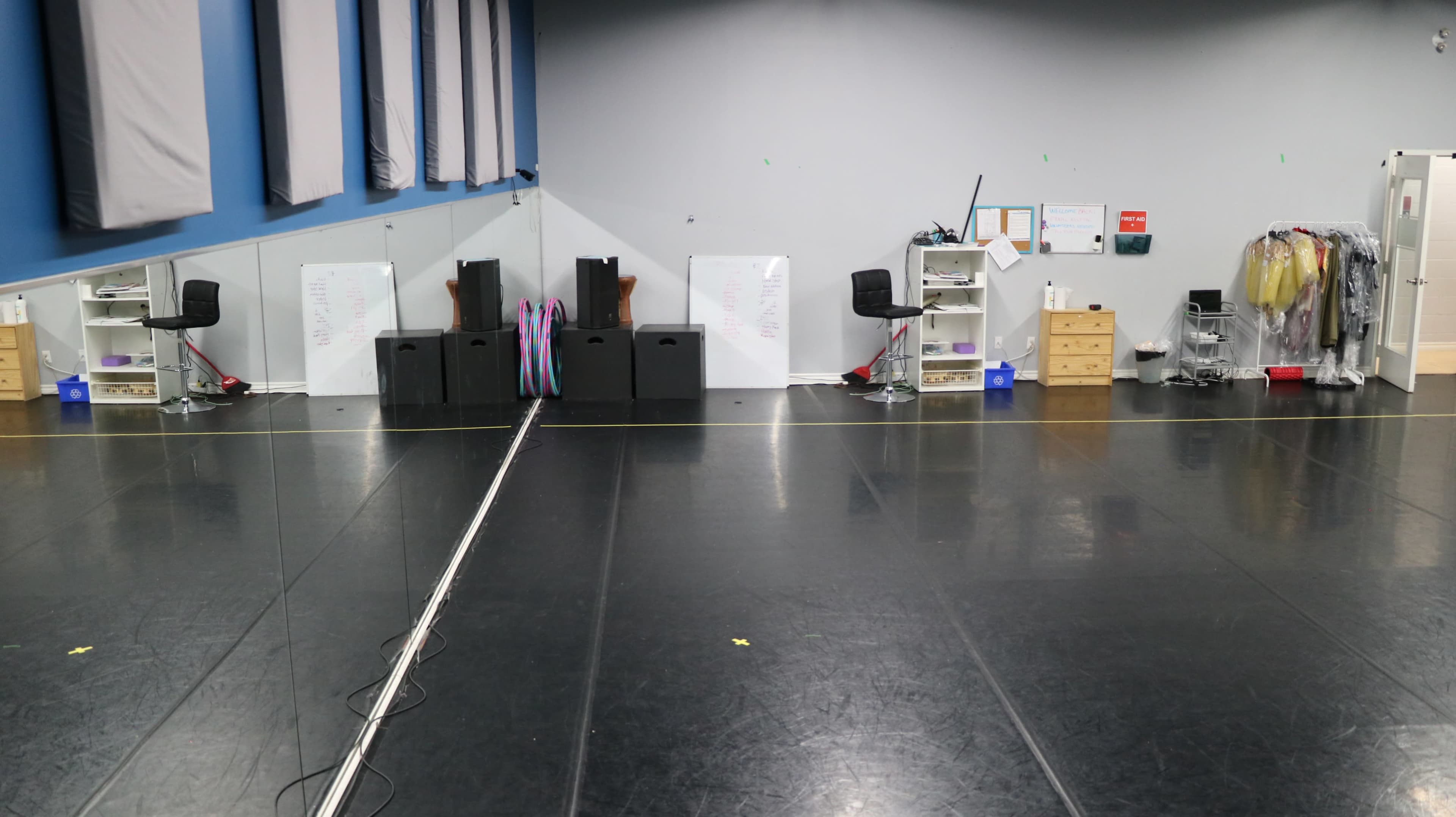 The image shows a dance studio with a mirrored wall, black flooring, and storage furniture on one side.