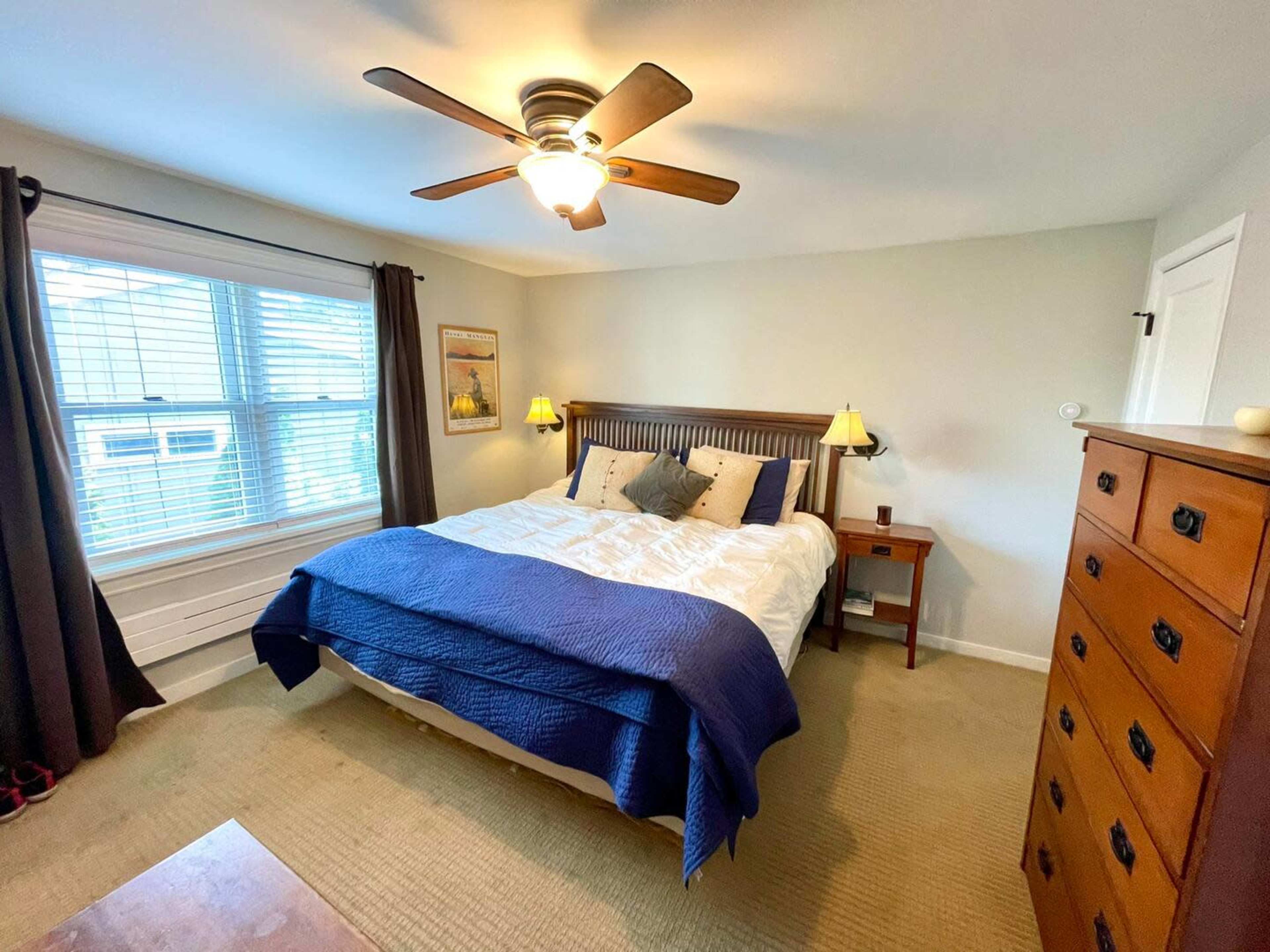 A neatly arranged bedroom featuring a bed with blue bedding, two bedside lamps, a wooden nightstand, and a dresser, all illuminated by ceiling light.