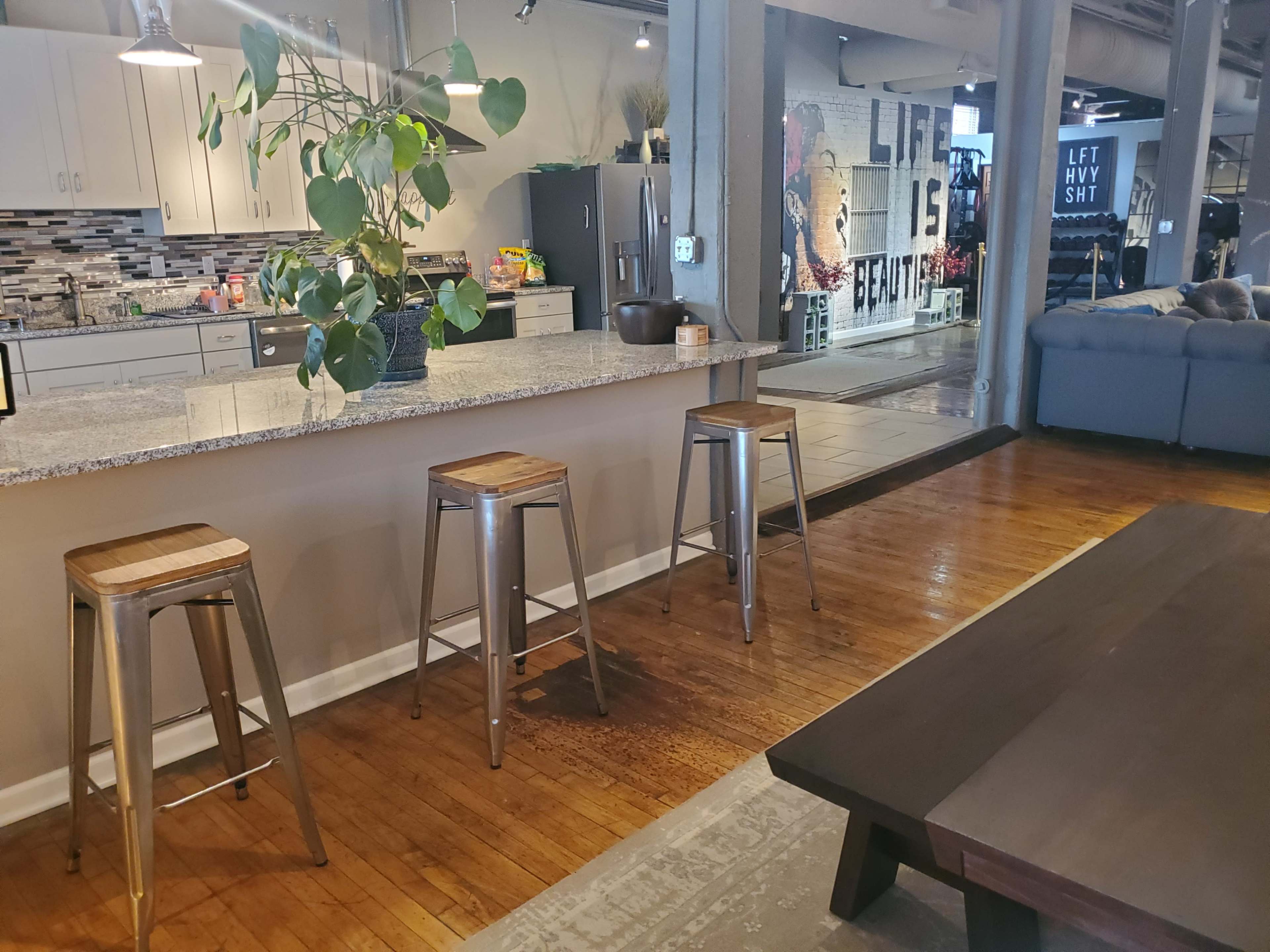 A modern kitchen area features a granite countertop with three metal bar stools and a vibrant wall mural in the background.