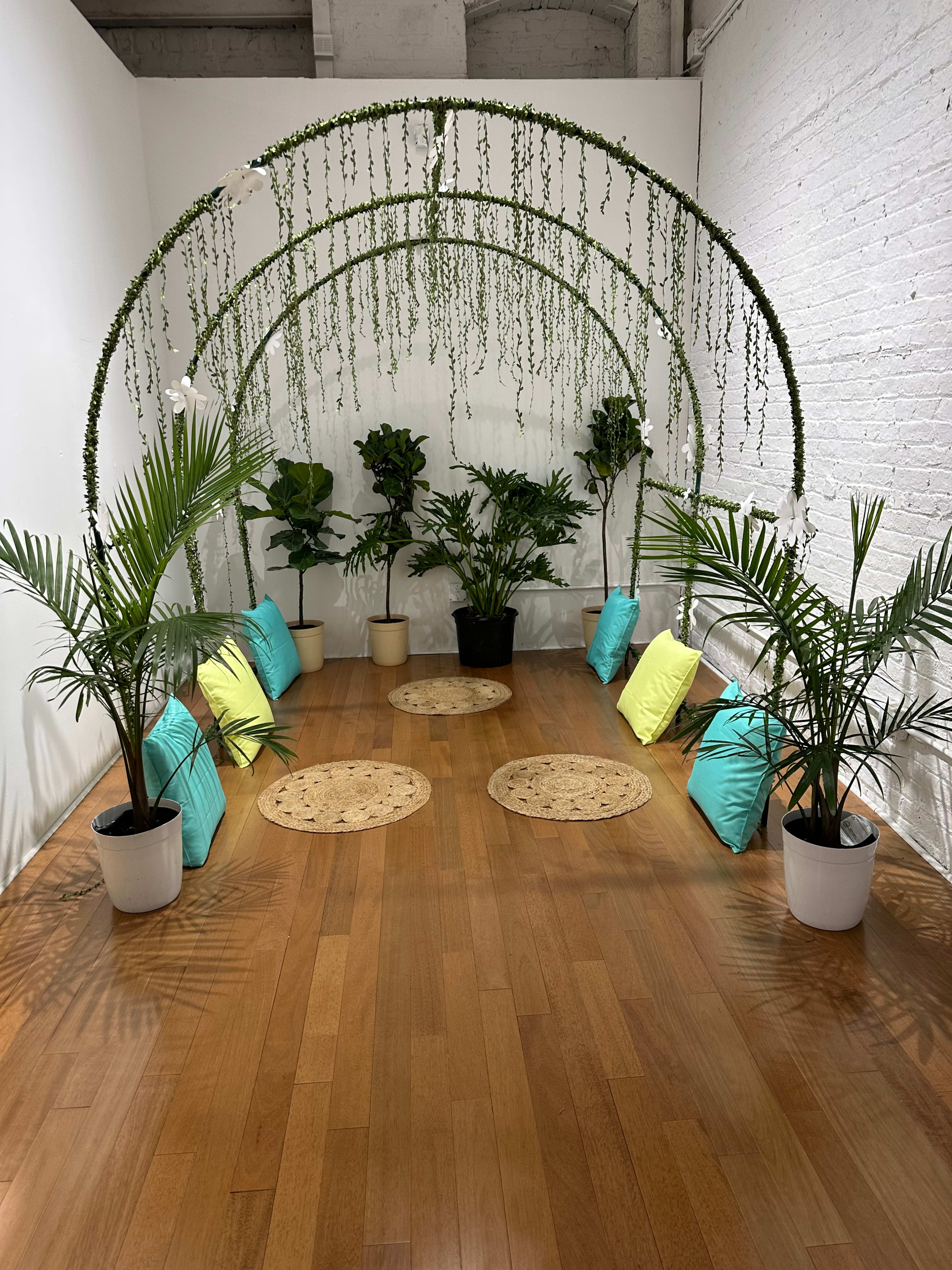 A small indoor space with a decorative arch covered in greenery, surrounded by potted plants and colored cushions arranged on a wooden floor.