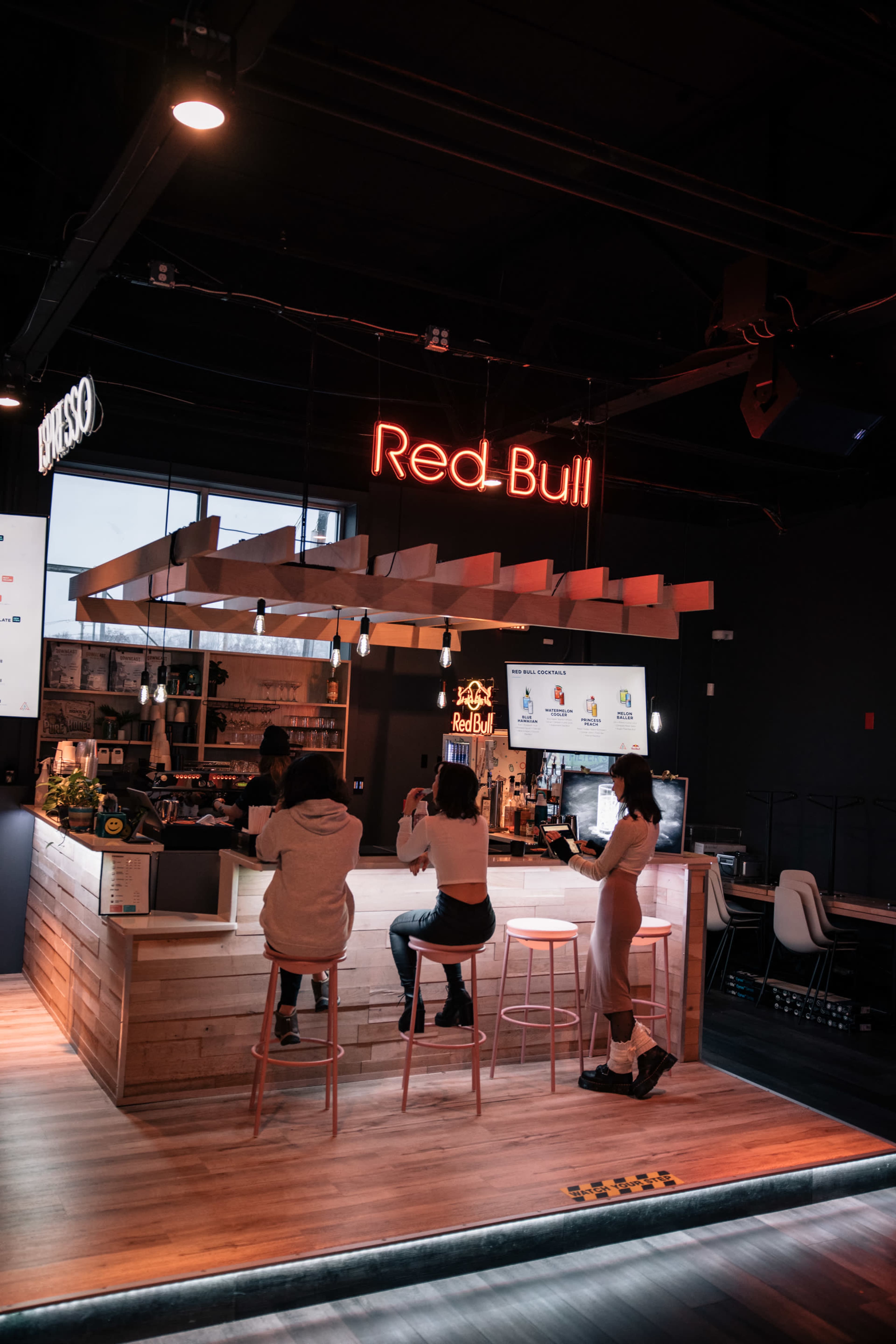 A modern bar area featuring a prominent "Red Bull" neon sign, where several people sit on high stools.