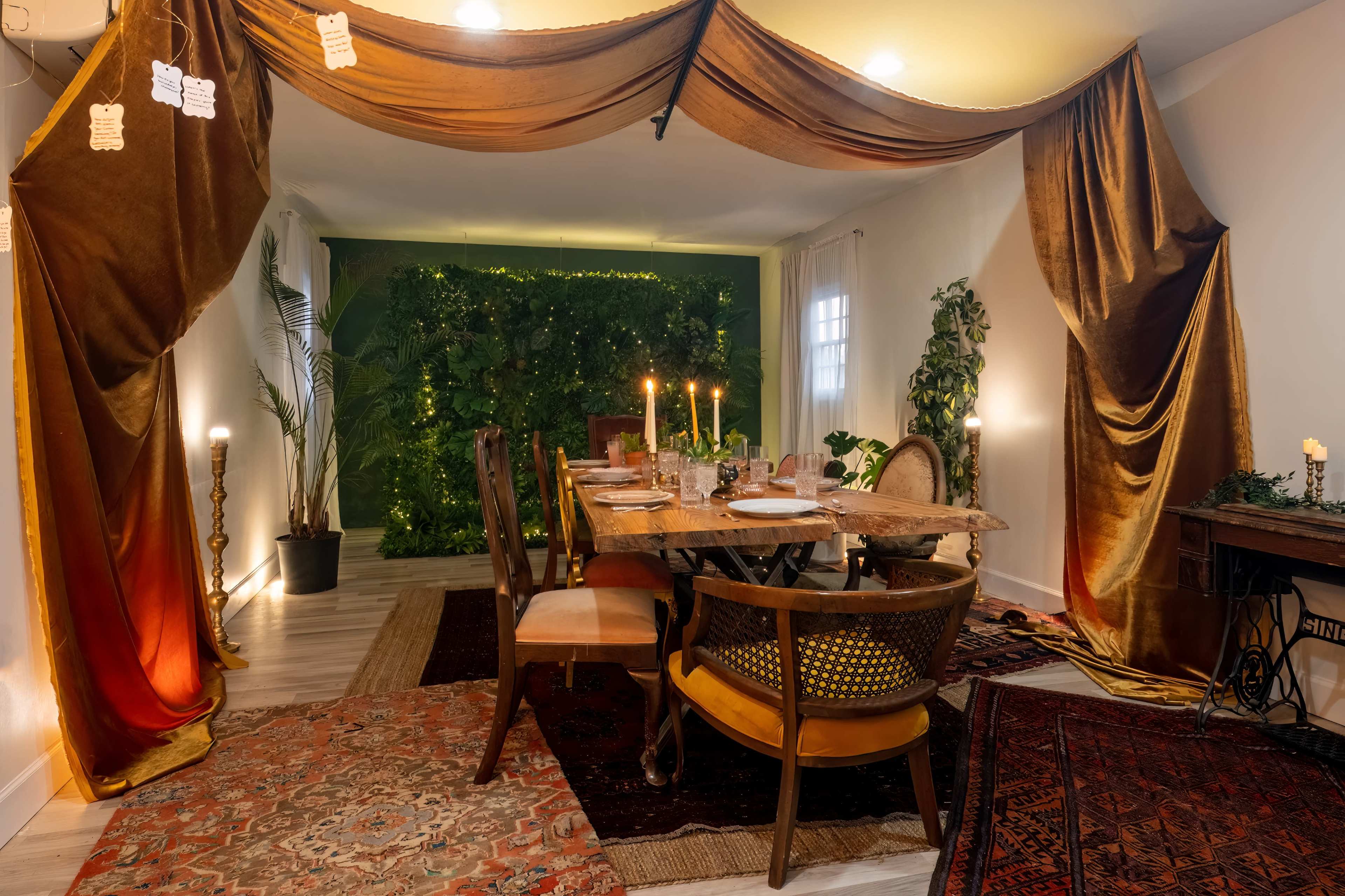 A dining area is set with a wooden table surrounded by chairs, accented by draped fabric and candles, with a green wall of plants in the background.