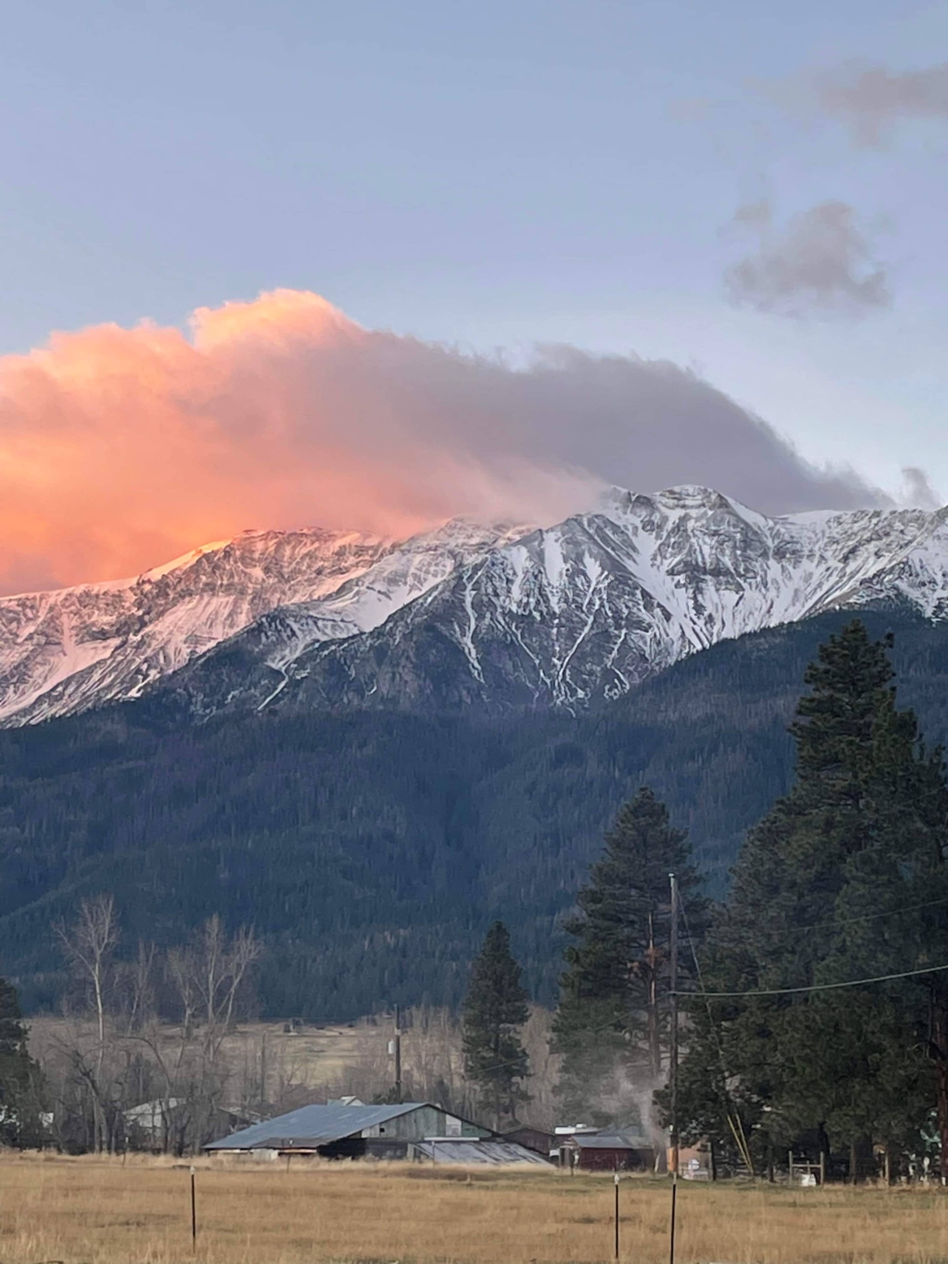 Snow-capped mountains rise in the background, partially obscured by clouds, with a grassy field and trees in the foreground.