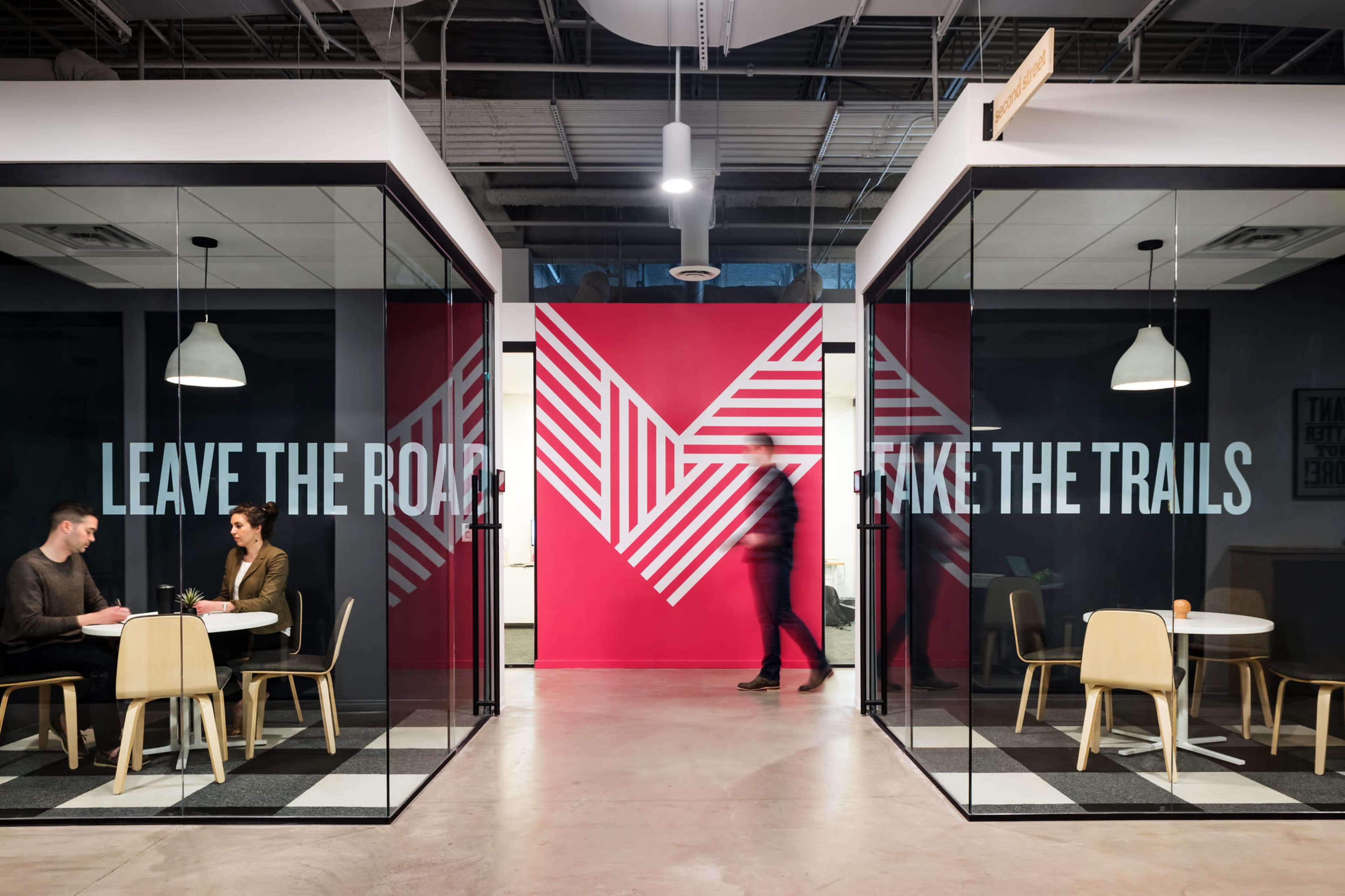 The image shows a modern office space featuring two glass-walled meeting rooms with large printed phrases, a colorful wall mural in the background, and minimalist furniture.