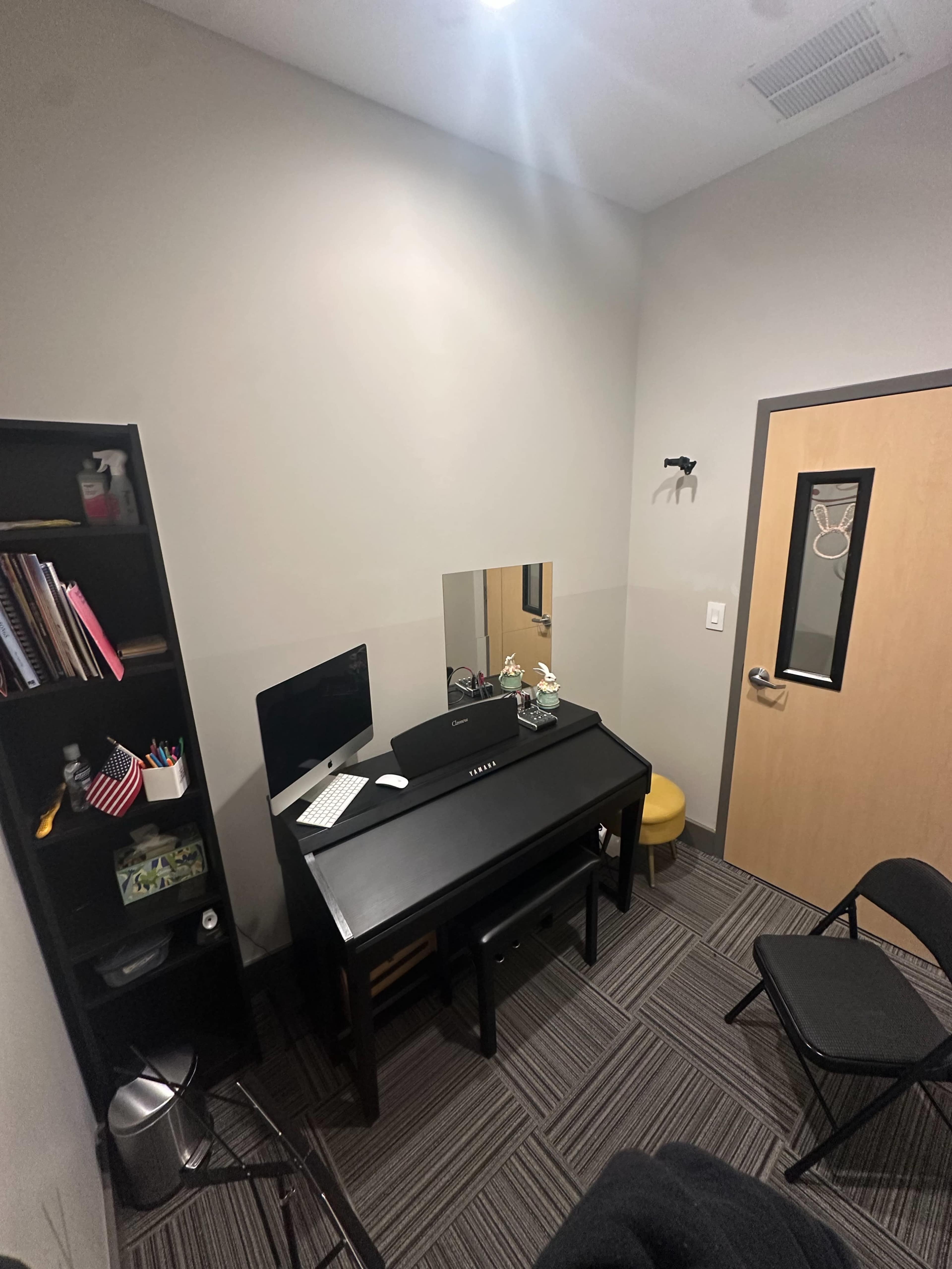 The image shows a small office or consultation room with a black desk, a mirror, a computer, and a chair, complemented by a bookshelf and a door with a glass window.