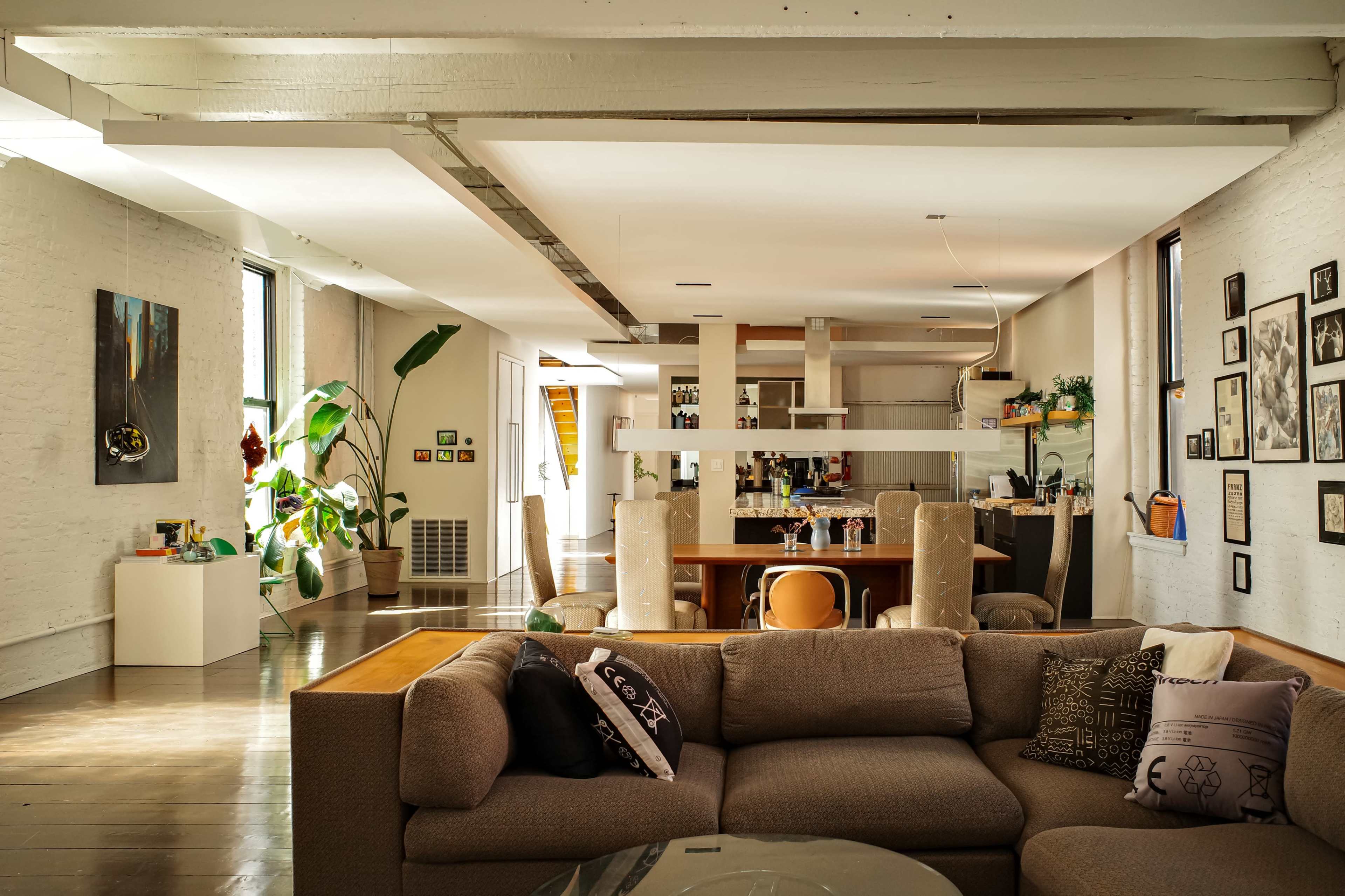 The image shows a spacious, modern living room with a sectional sofa, large windows, and an adjoining dining area featuring a kitchen in the background.