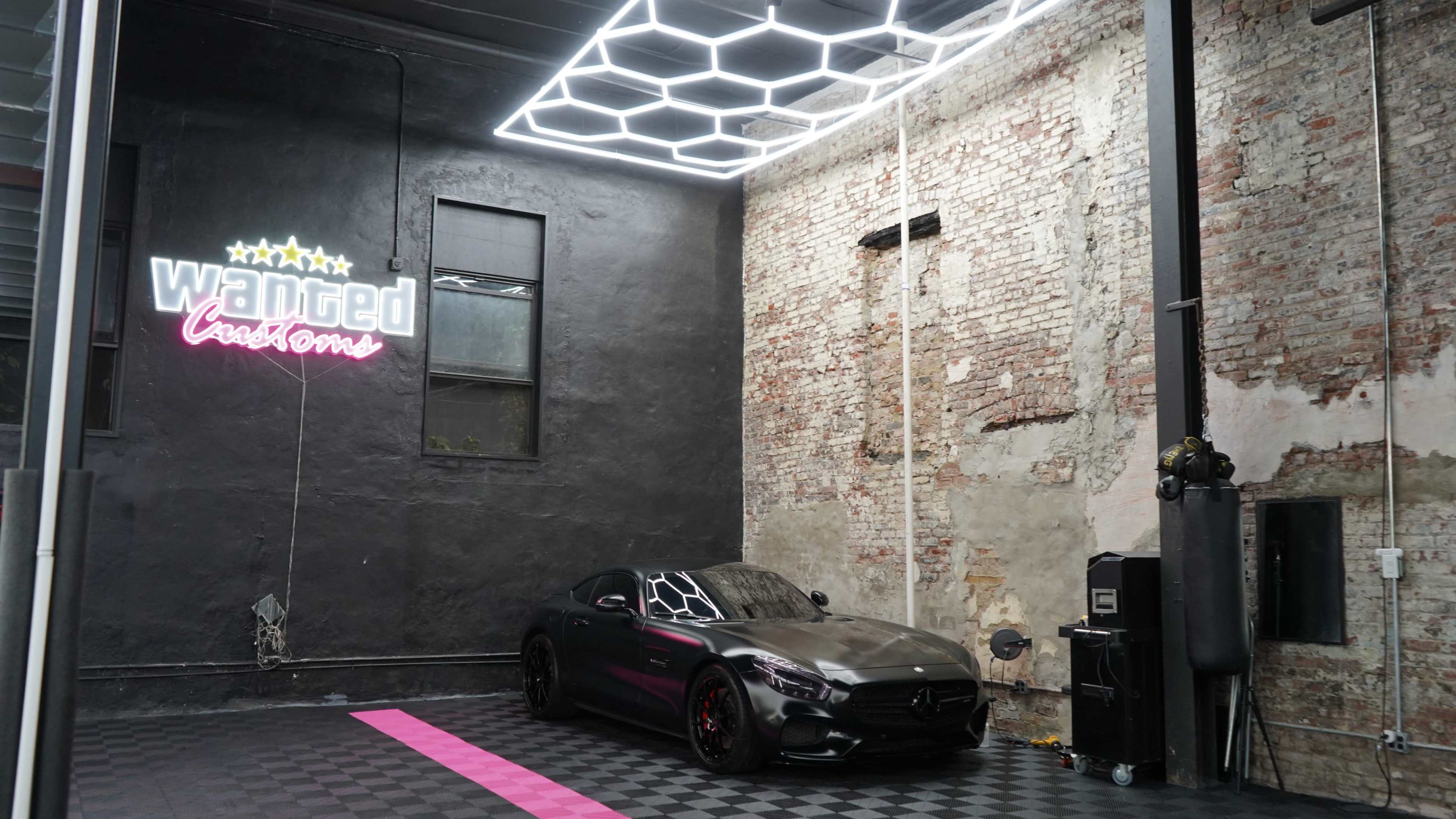 The image shows a sleek black Mercedes-Benz sports car parked in a brightly lit garage with exposed brick walls and modern lighting.
