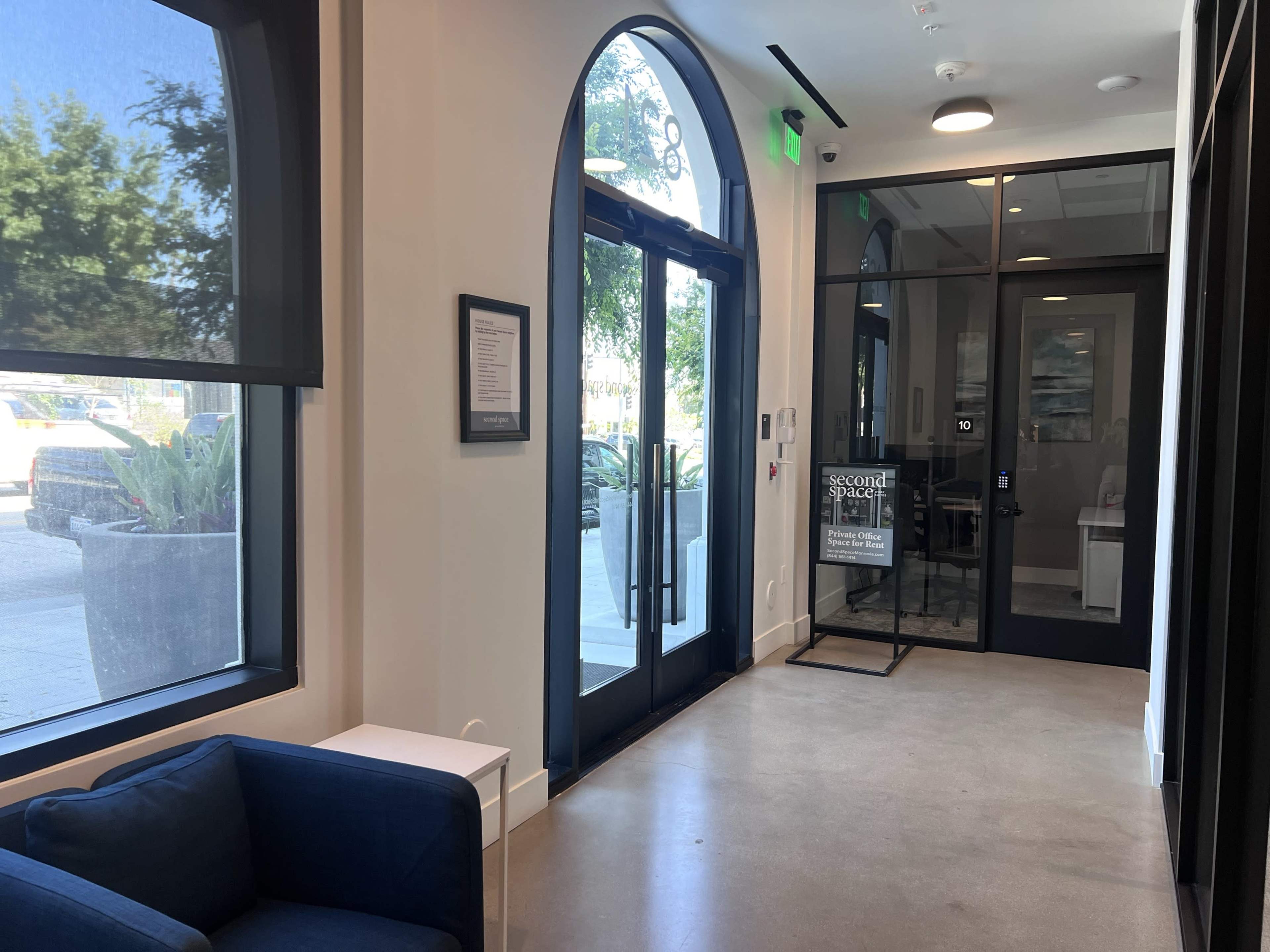The image shows a contemporary hallway with a blue couch, large arched windows, and glass doors leading to an exterior area.