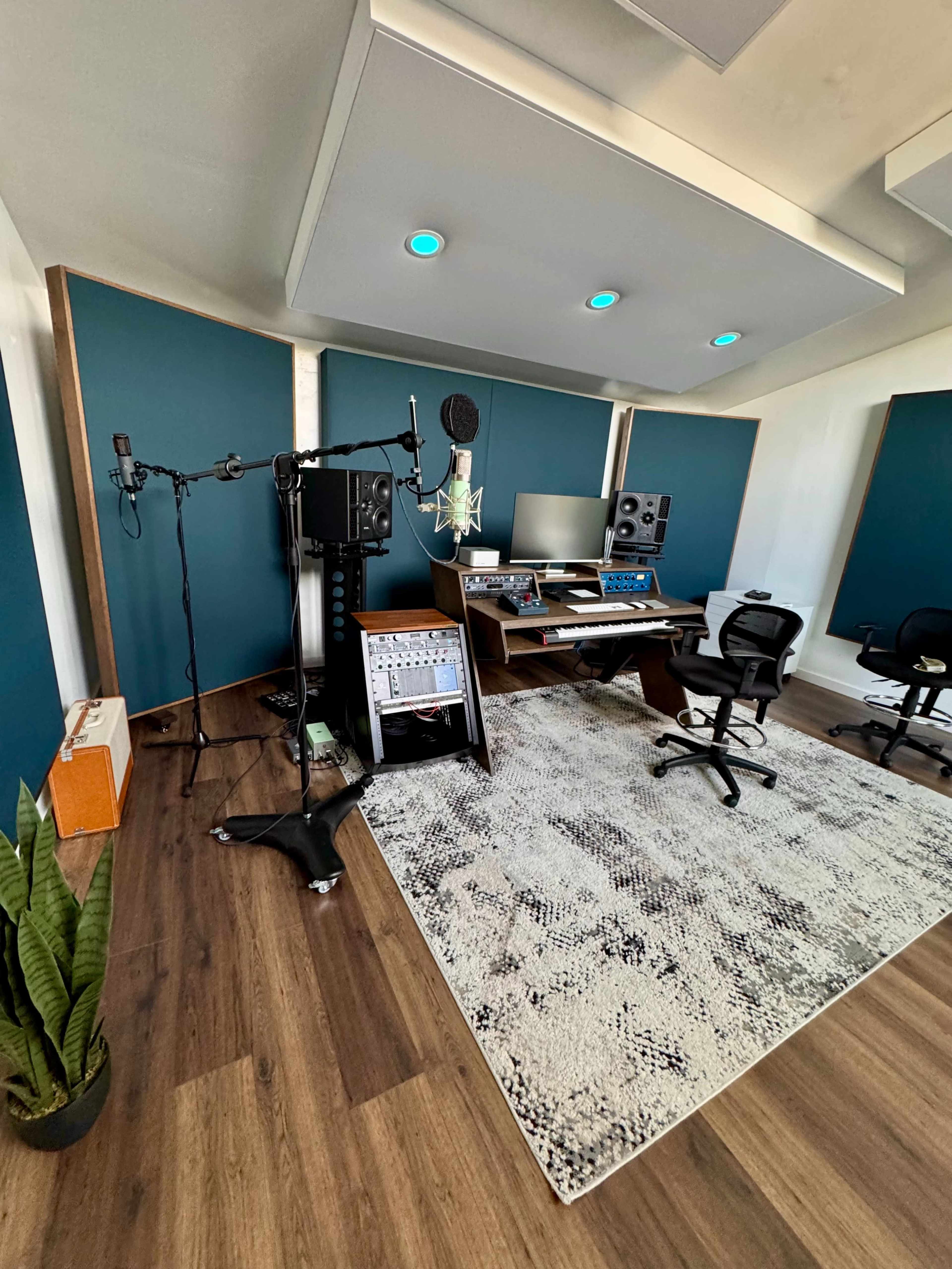 The image shows a modern recording studio with soundproof panels, a mixing console, microphones on stands, and comfortable chairs on a textured rug.
