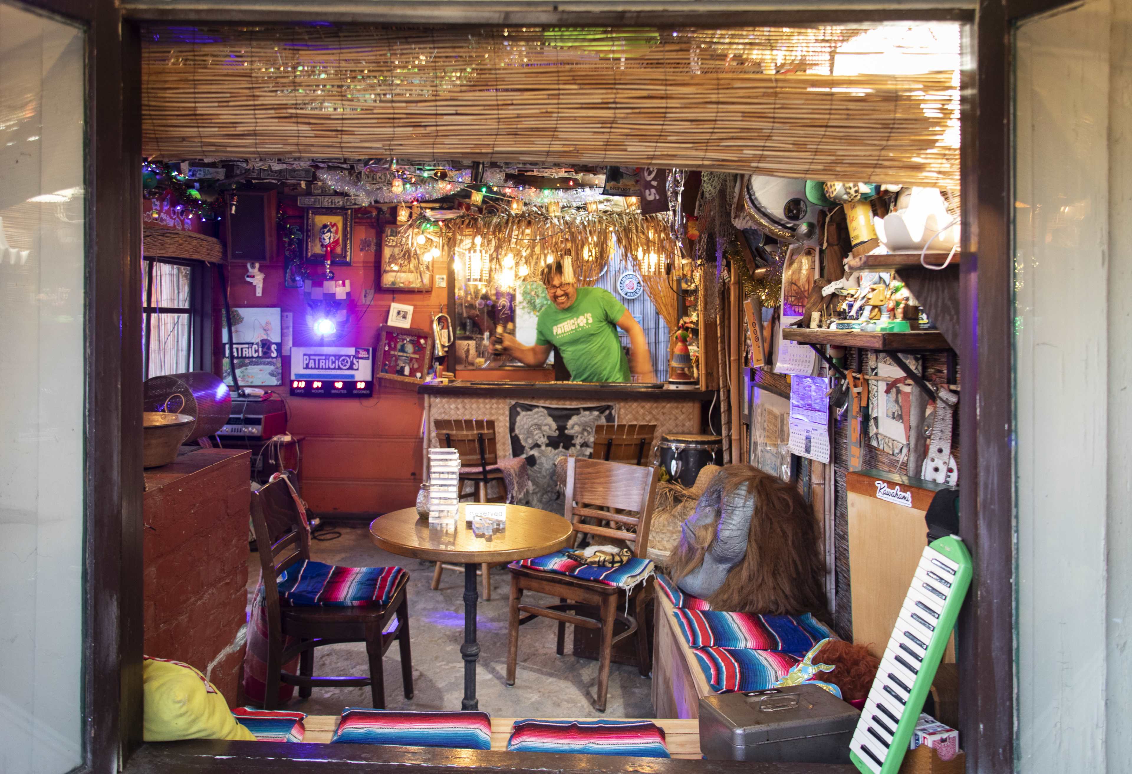 A cozy room features colorful seating, various decorations, and a bar area visible through an open window.