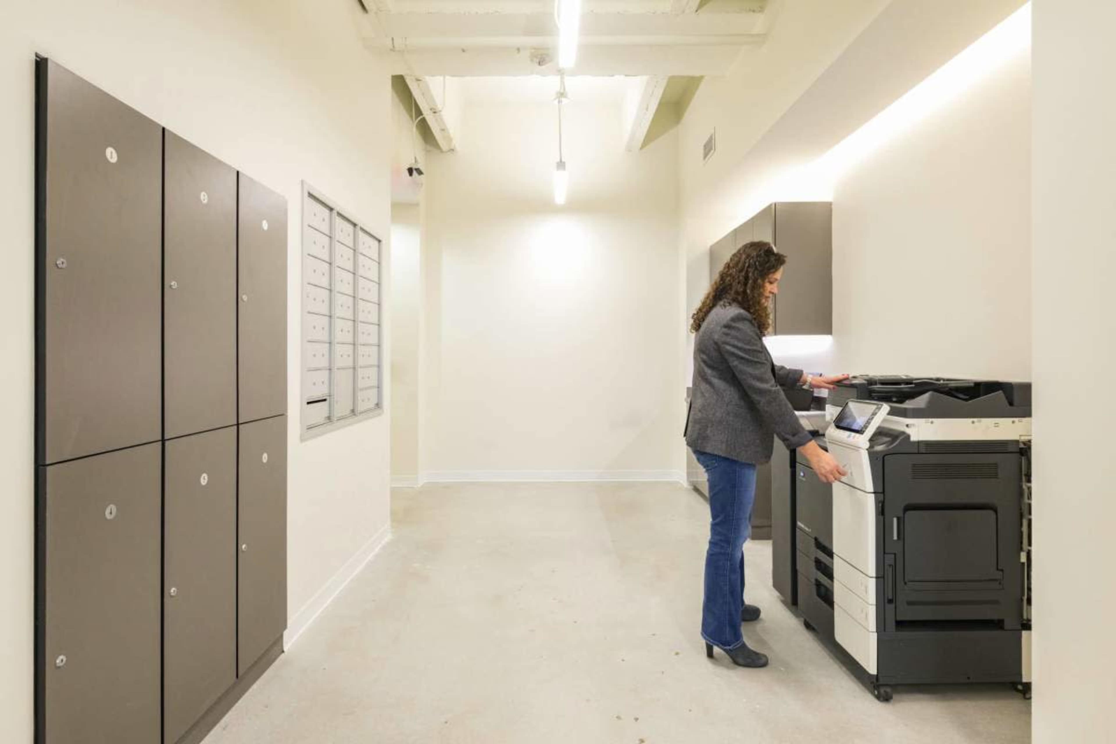 A woman is using a multifunction printer in a well-lit hallway with mail lockers and storage cabinets.
