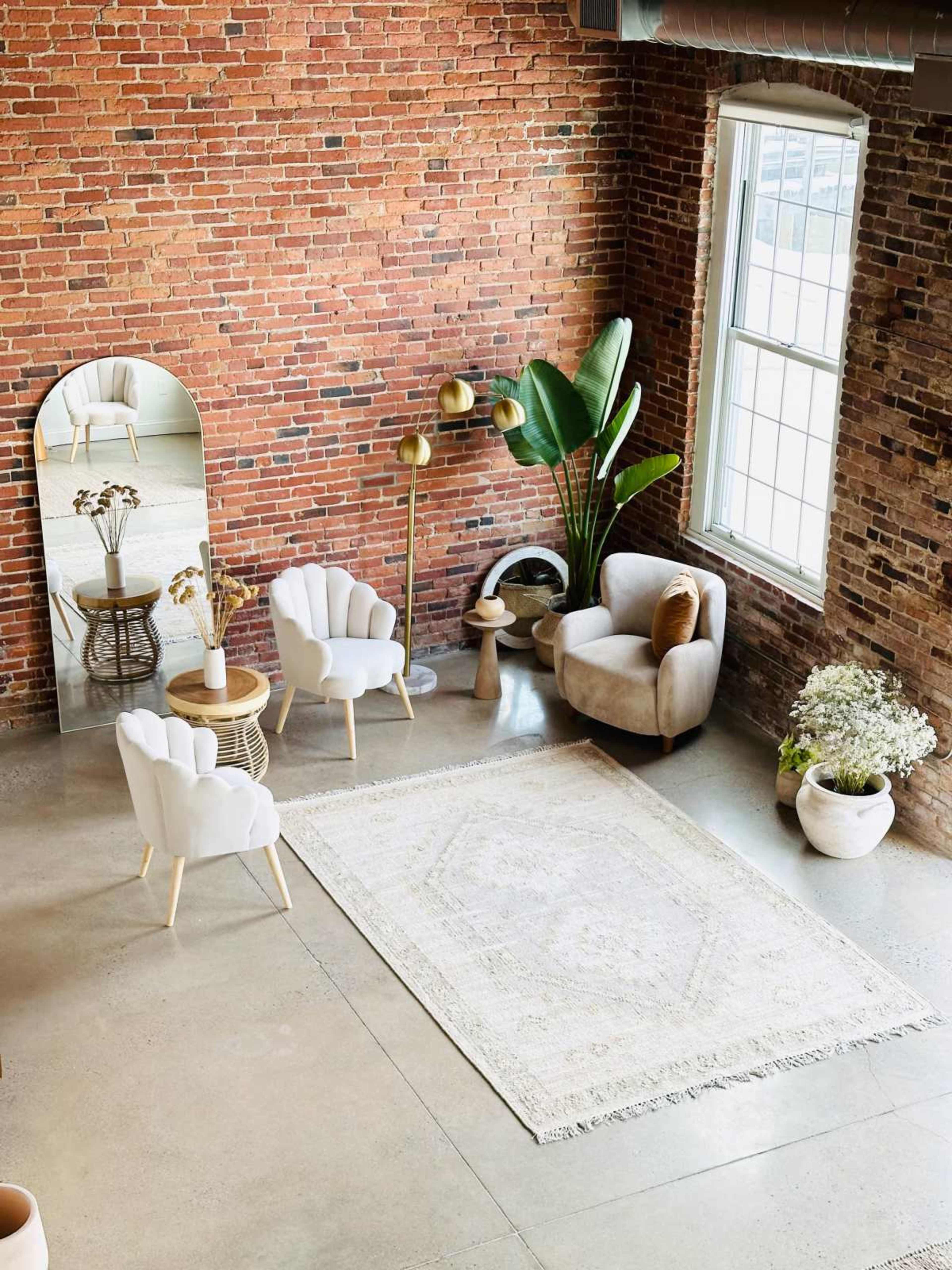 The image shows a cozy living area with a brick wall, two white chairs, a beige armchair, a round coffee table, a large mirror, a gold floor lamp, a potted plant, and a decorative area rug.