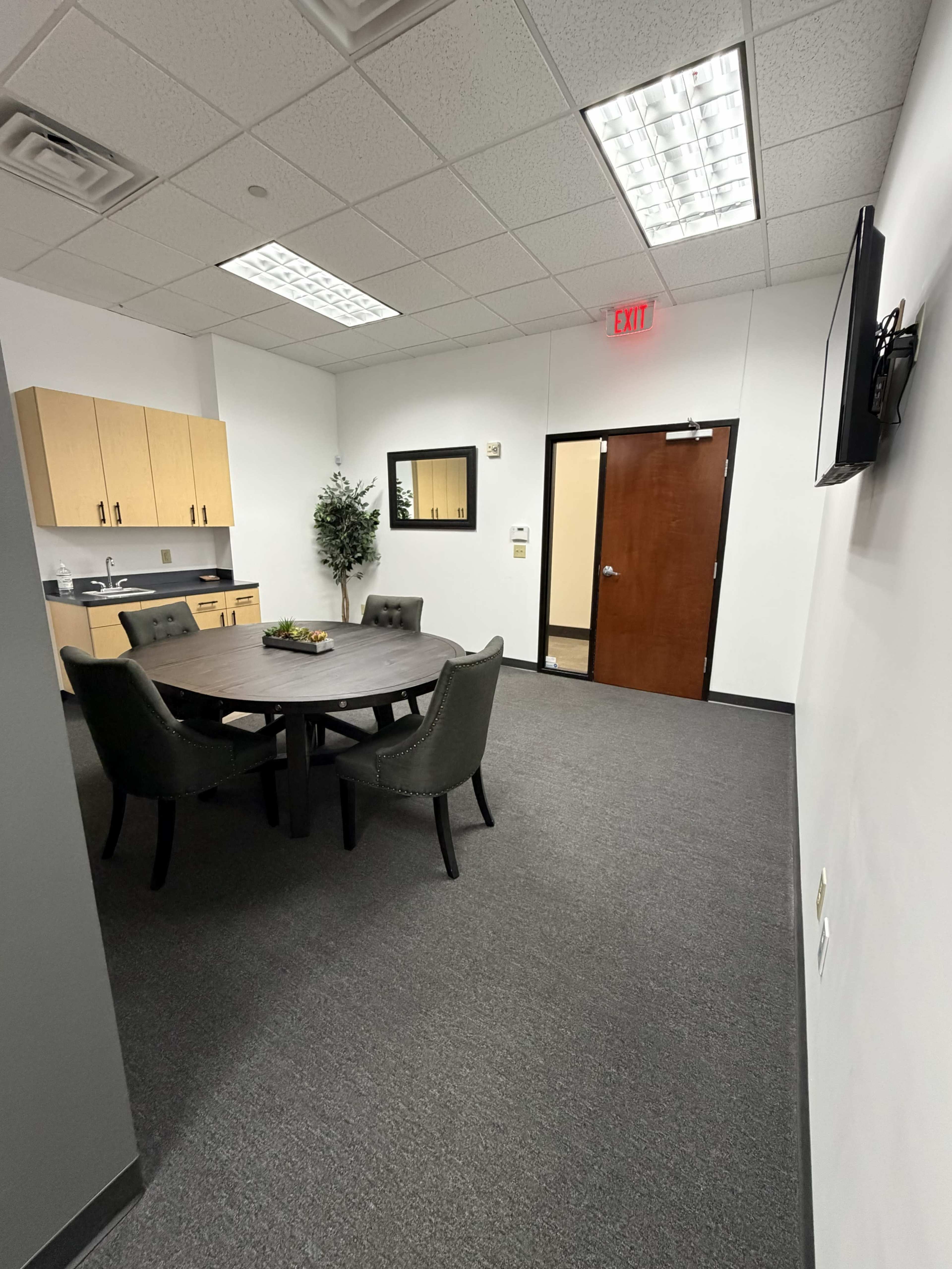 The image shows a small meeting room featuring a round table with four chairs, a kitchenette area with cabinets, and a wall-mounted television.