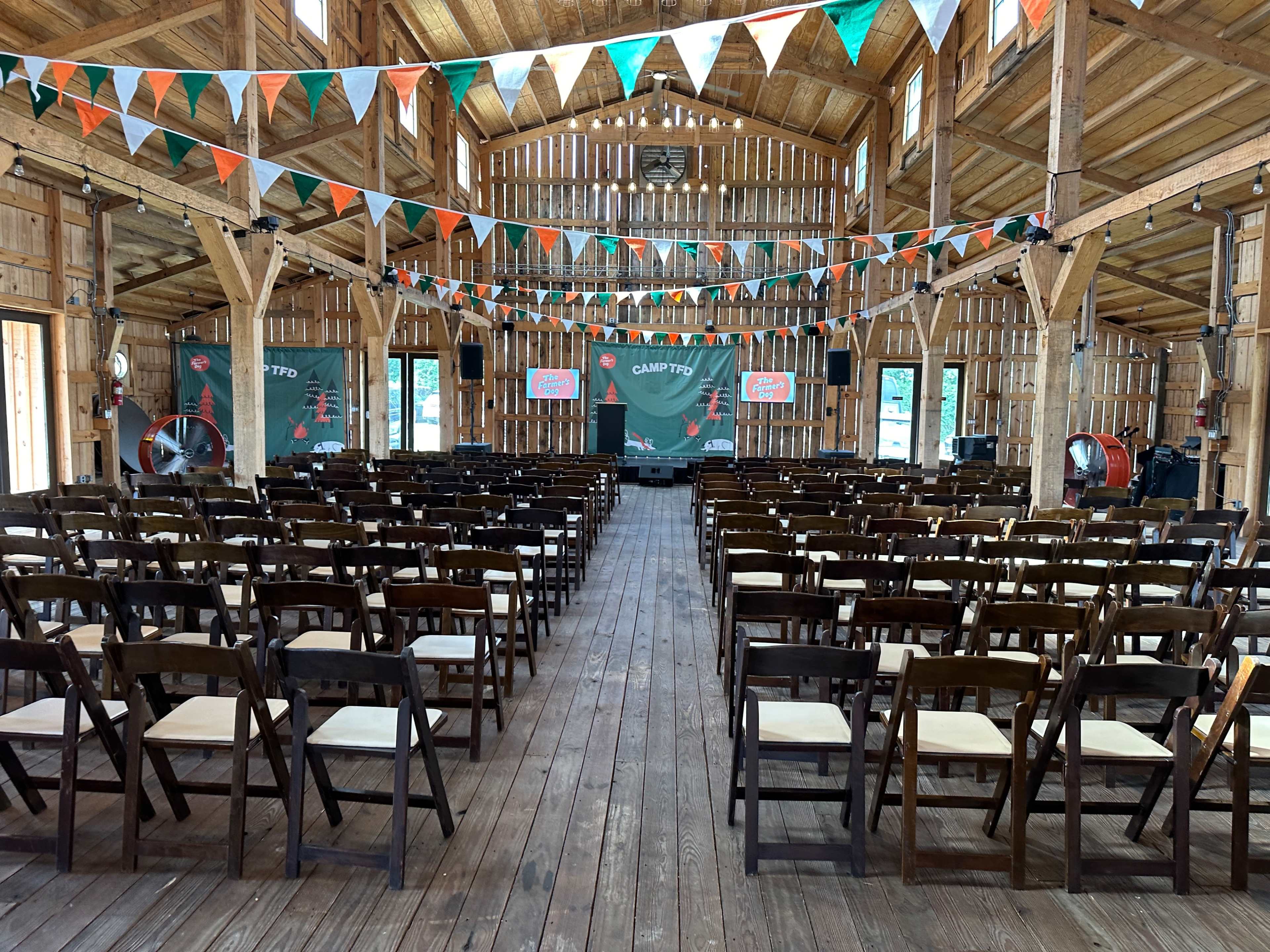 The image shows the interior of a wooden barn decorated with colorful banners and arranged with rows of wooden chairs facing a stage.