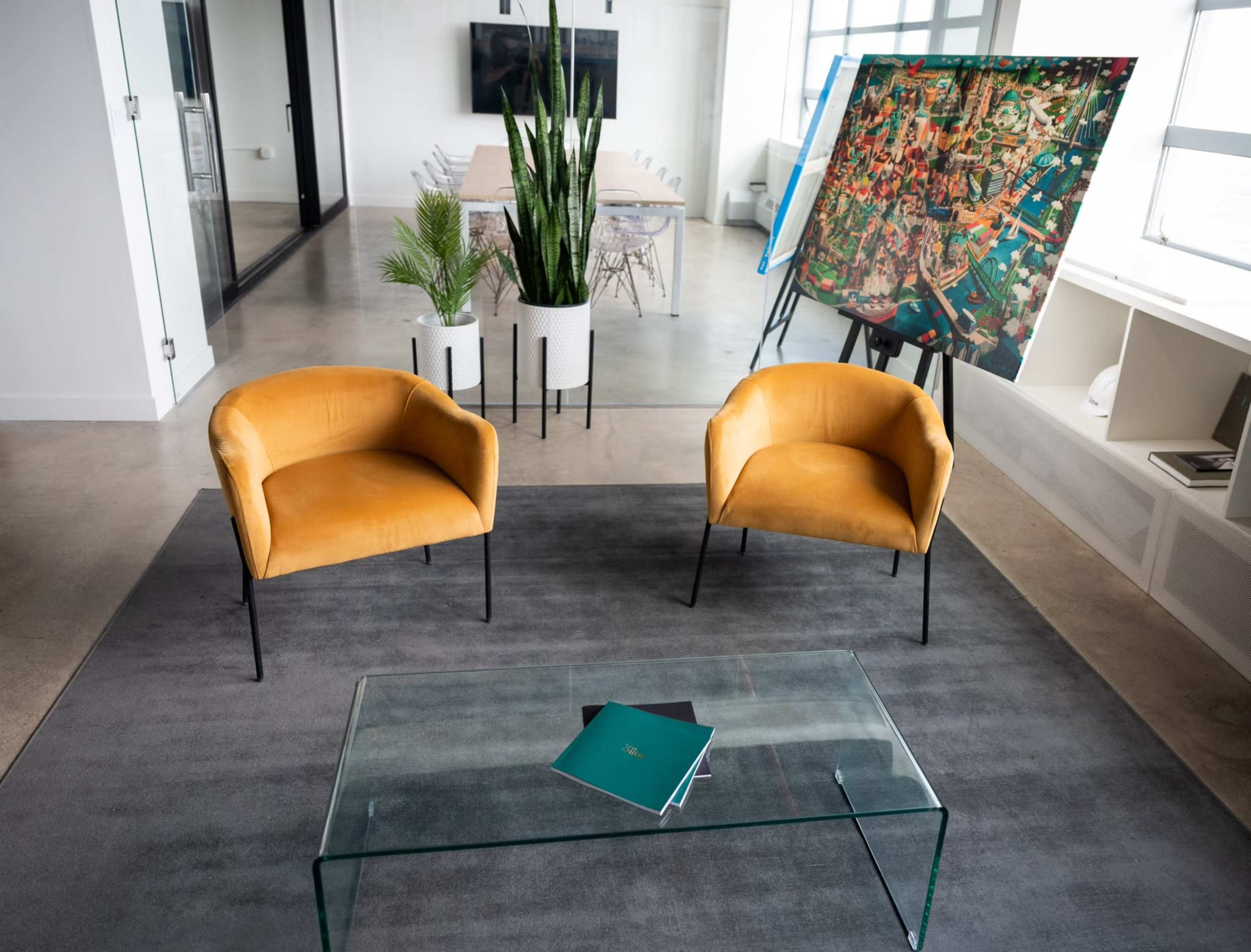 Two yellow chairs are positioned around a glass coffee table in a modern office space with a large artwork and potted plants in the background.