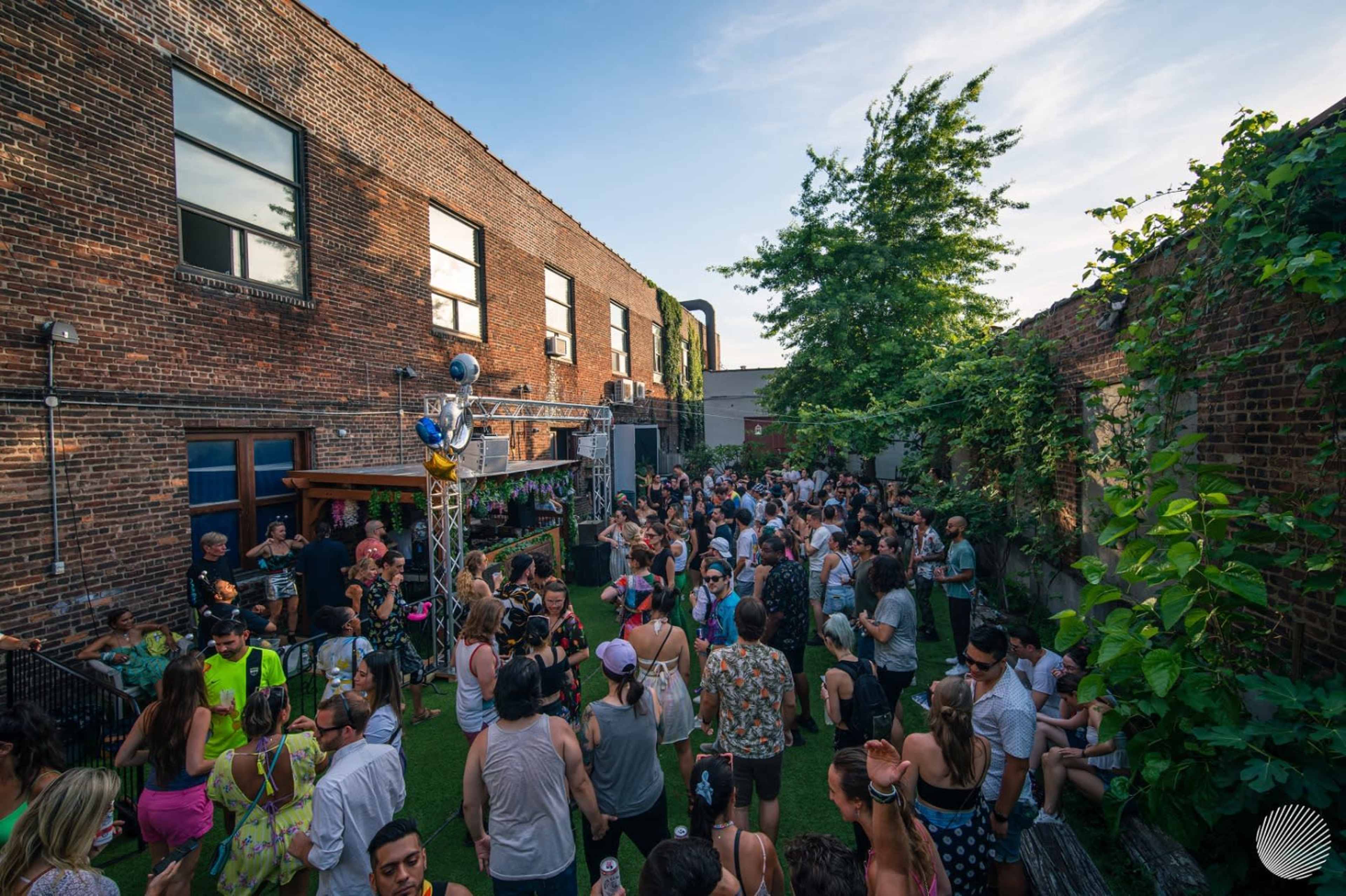 A large crowd of people gathers in a vibrant outdoor space surrounded by brick walls and greenery, enjoying a lively event.