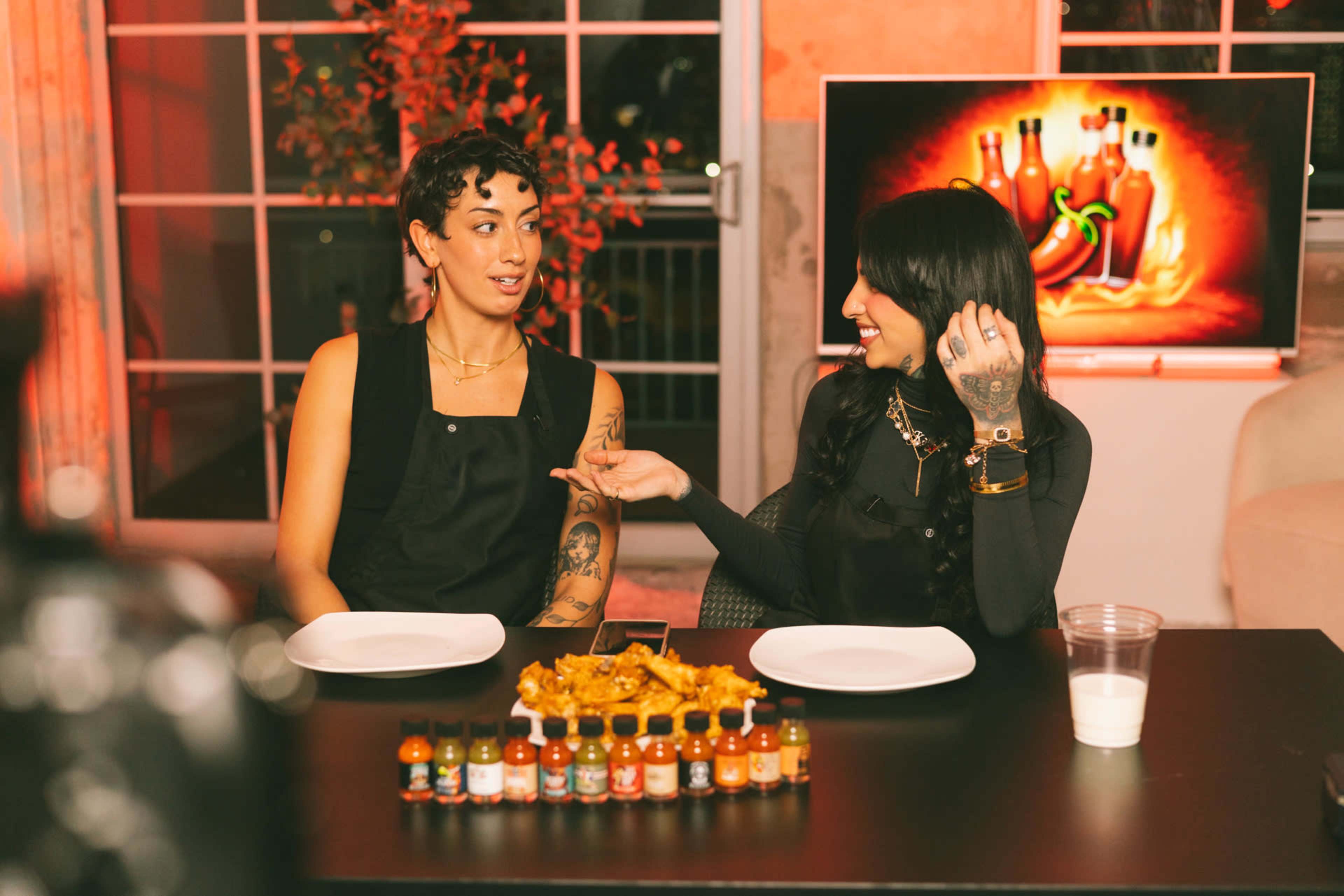 Two individuals are seated at a table set with empty plates and a collection of hot sauce bottles, with a colorful visual display of chili peppers in the background.