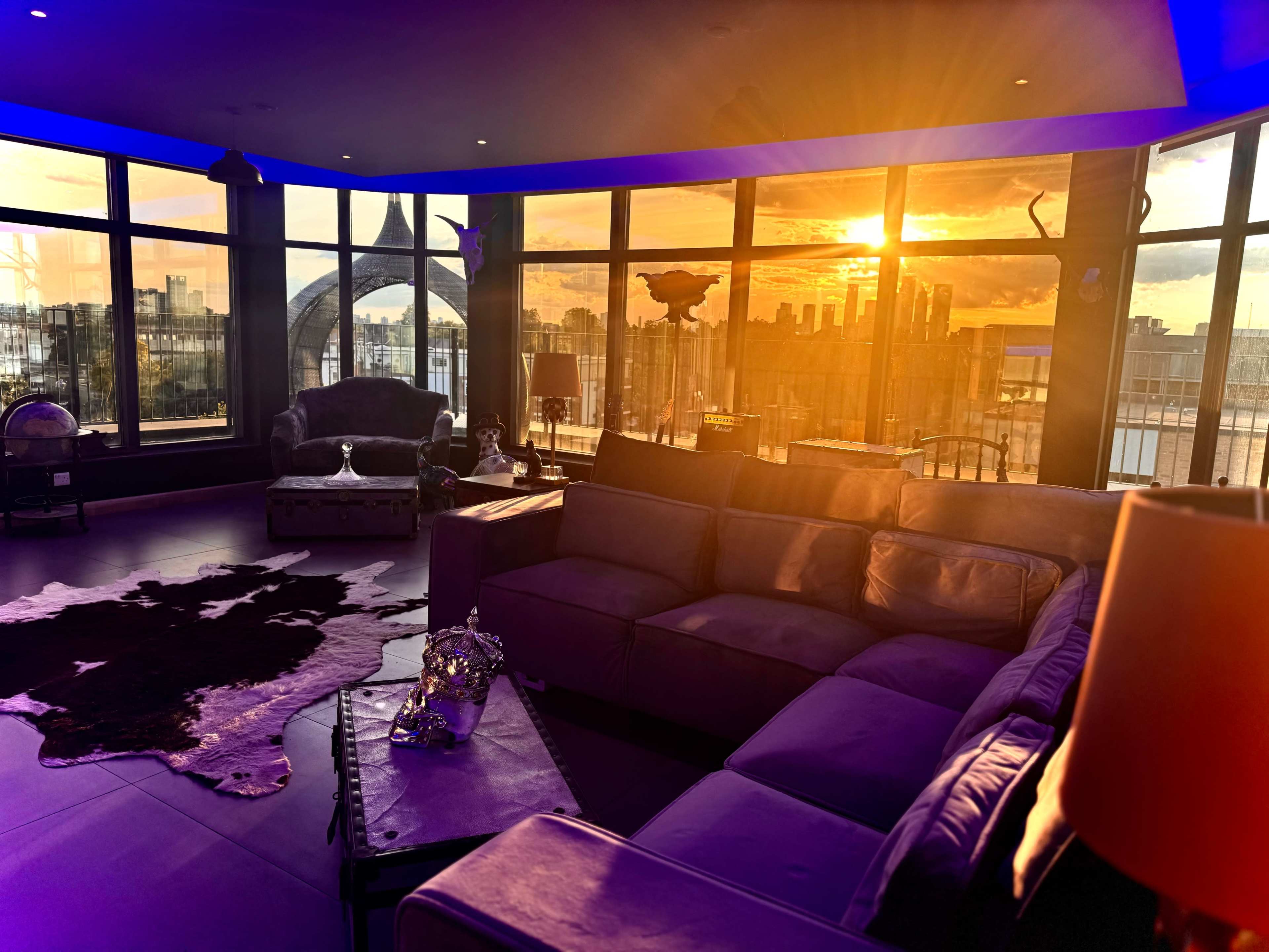 A modern living room features a sectional sofa, large windows displaying a sunset over a city skyline, and decorative accents including a cowhide rug.