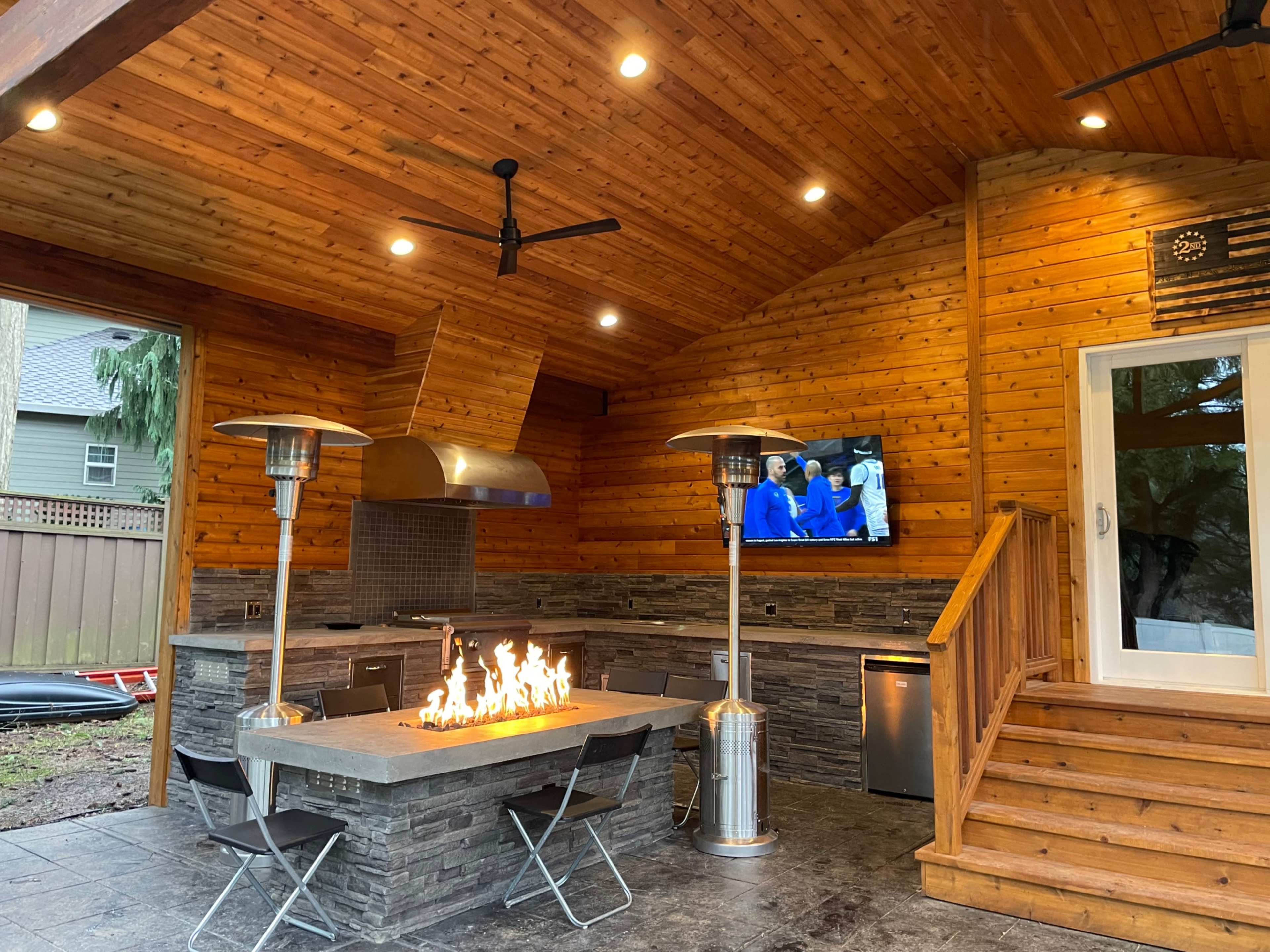 A modern outdoor kitchen with a stone countertop, a built-in grill, two patio heaters, and a television mounted on the wooden wall.