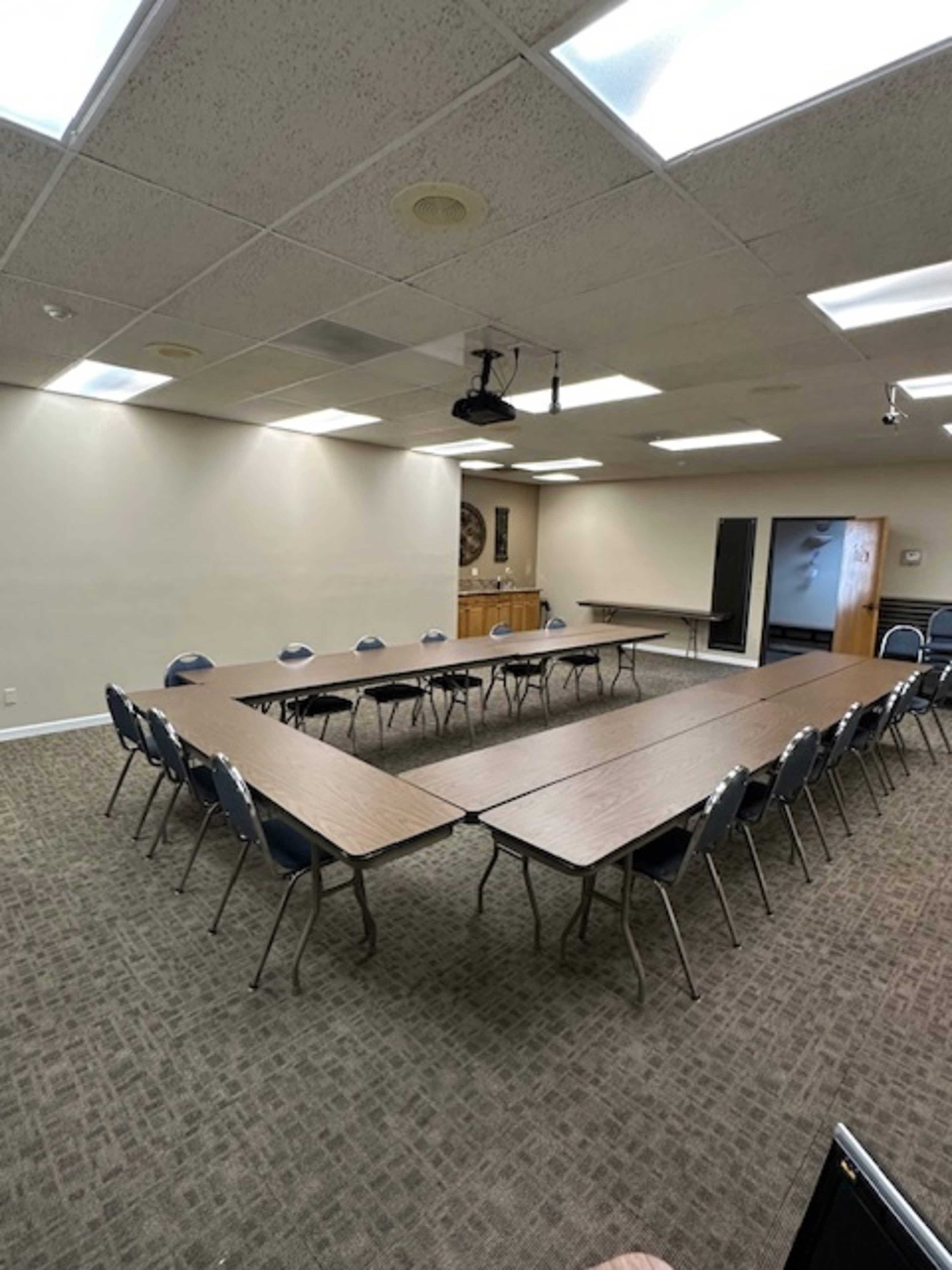 A conference room with two long tables arranged in a U-shape and chairs placed around them, featuring a projector overhead.
