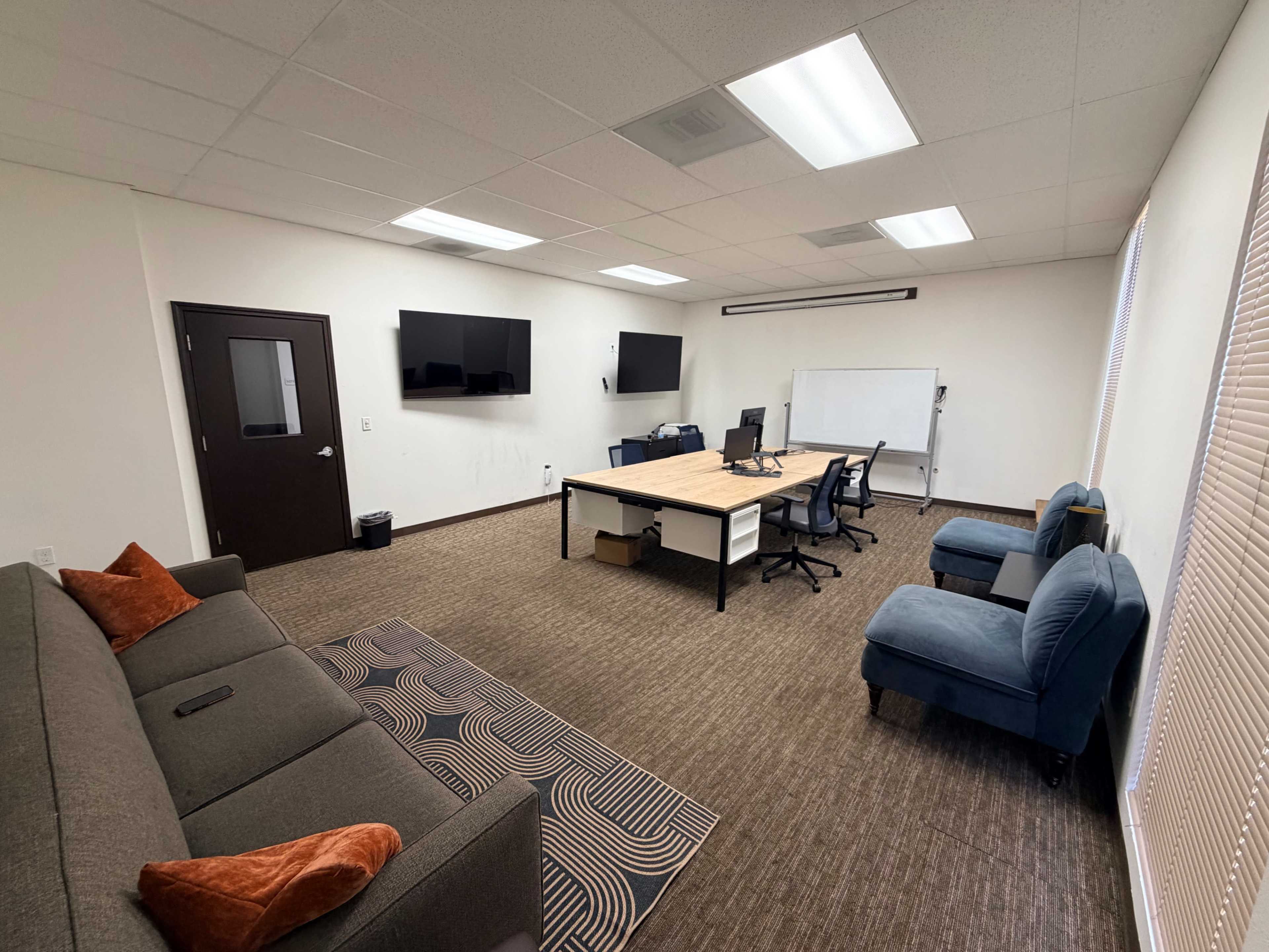 The image shows a conference room featuring a large table with chairs, a couch, two wall-mounted screens, and a whiteboard.
